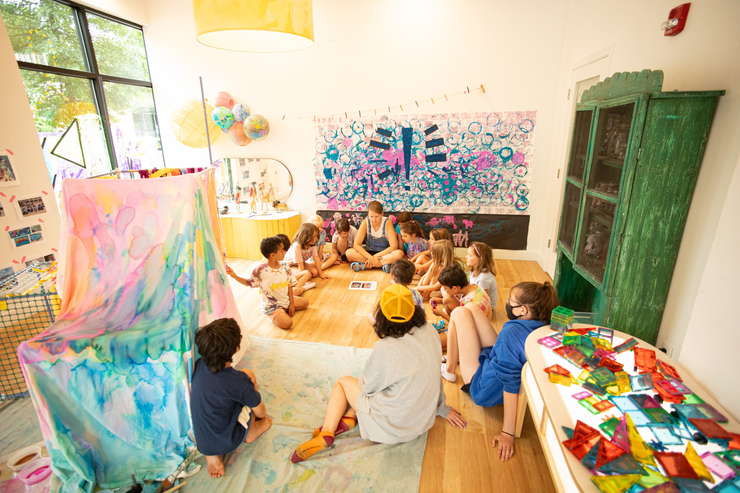 Children and an adult sitting in a circle on the floor in an art classroom, with colorful artwork, craft supplies, and decorations surrounding them.