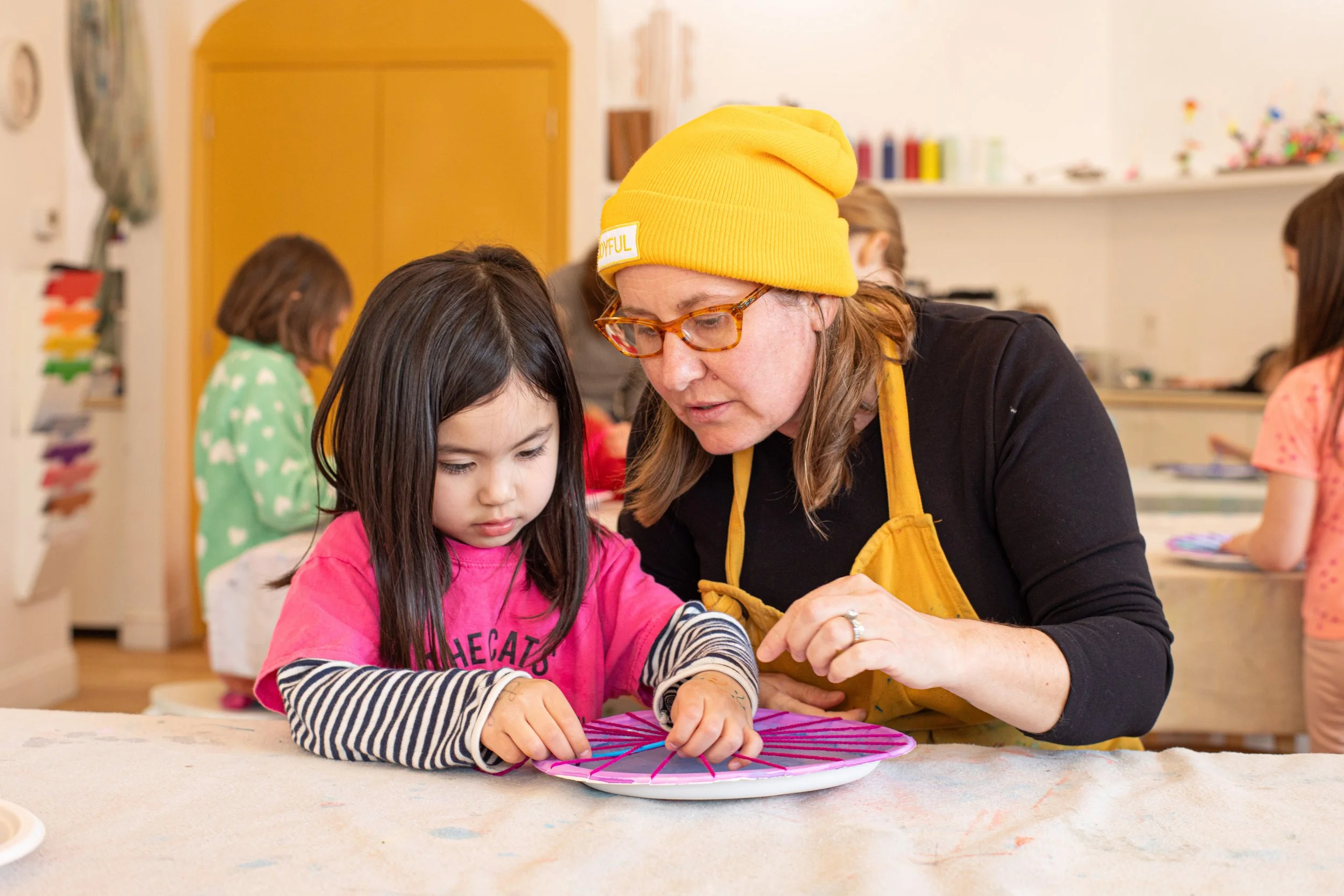A young girl being guided by an adult woman in a yellow beanie and apron, as they work on an art project in a classroom setting with other children in the background.