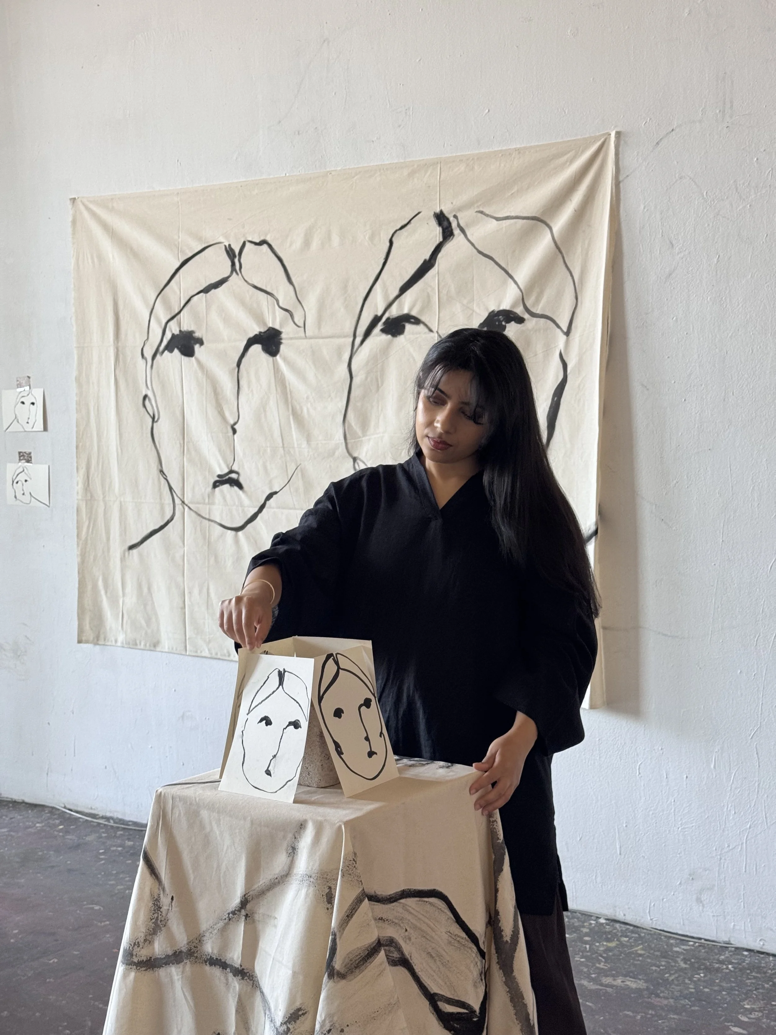 Artist girl working in her studio, standing in front of a fabric banner with a line drawing of faces, with additional paper artwork depicting faces on a table and hanging on the wall.