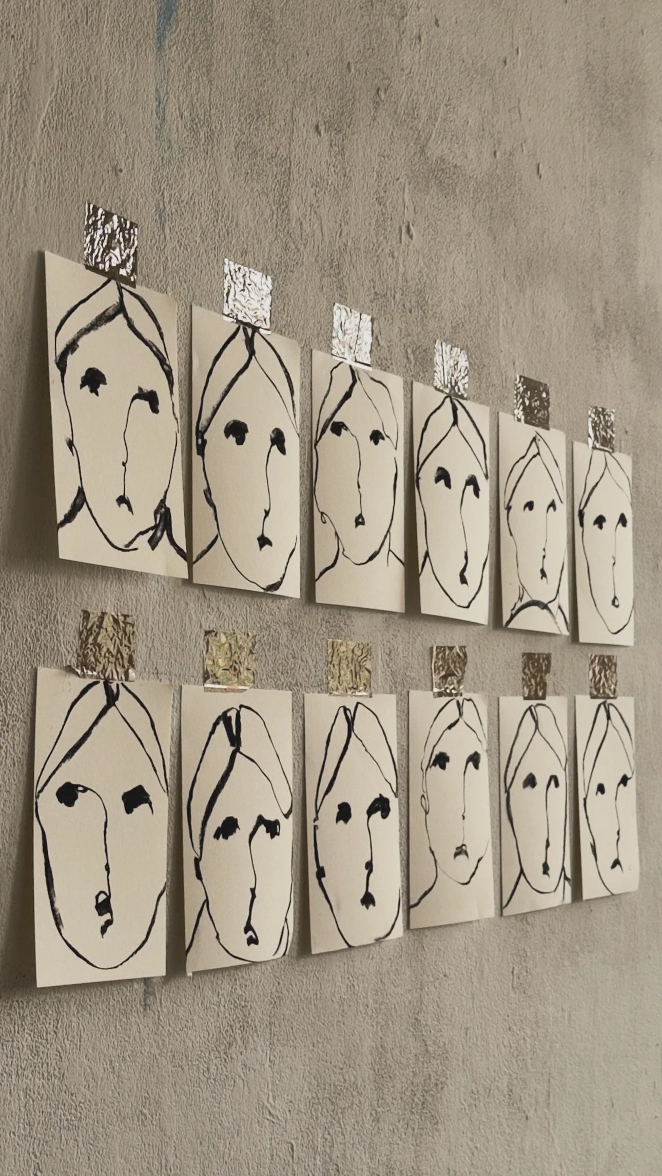 A collection of black and white abstract portrait sketches of faces on white paper, arranged in two rows on a textured wall, with shiny square pieces of metallic foil attached above some of the portraits.