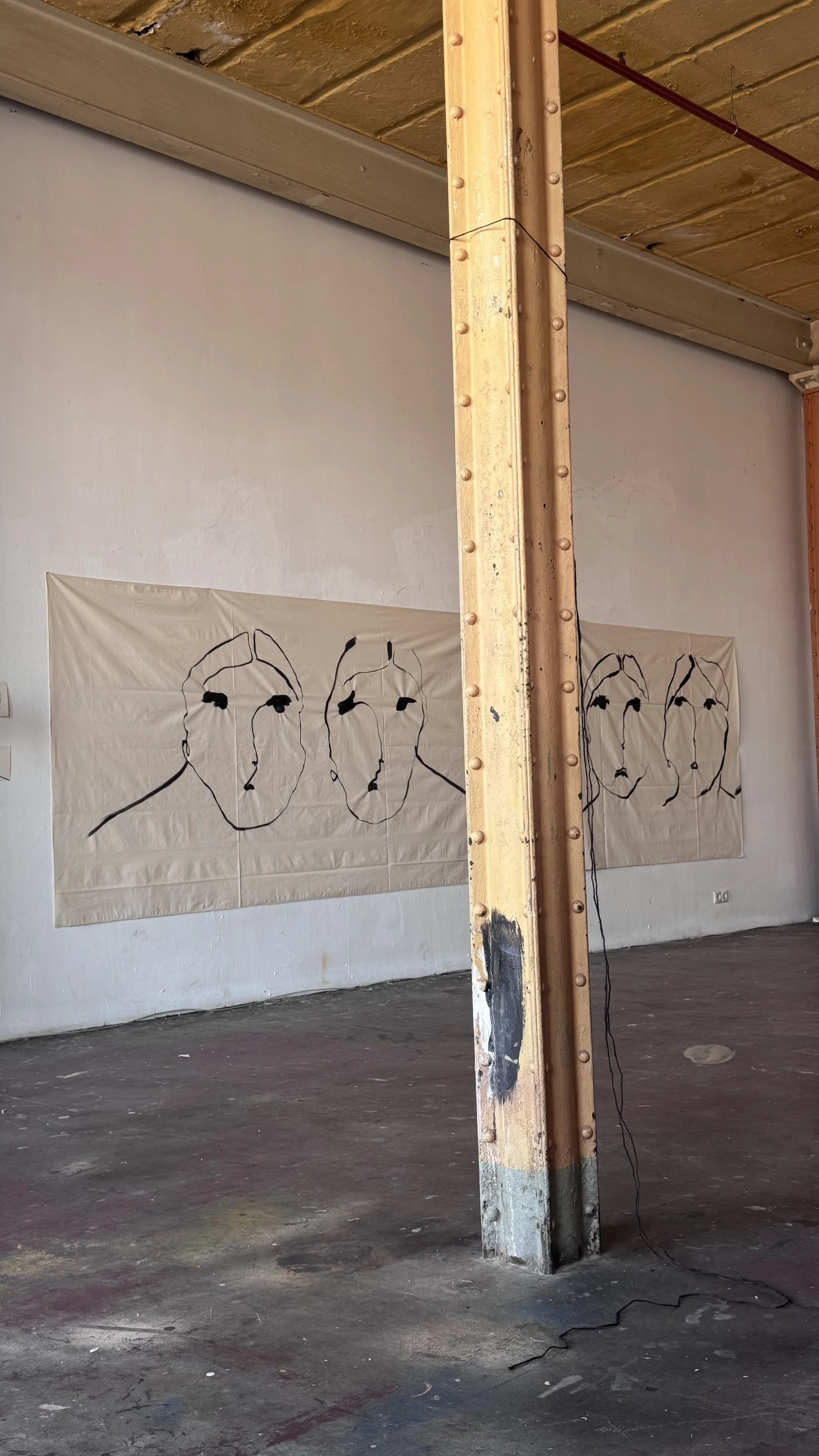 Art installation featuring three large, minimalist line drawings of human faces on a white cloth hanging on a wall in an industrial-style space.