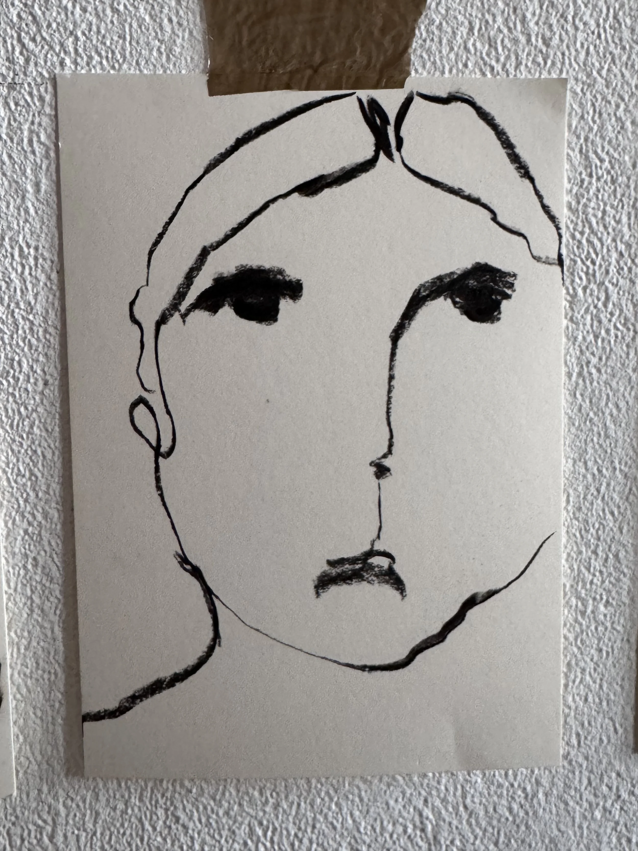 A black-and-white line drawing of a person's face, with bold, expressive features, on a piece of paper taped to textured white wall.