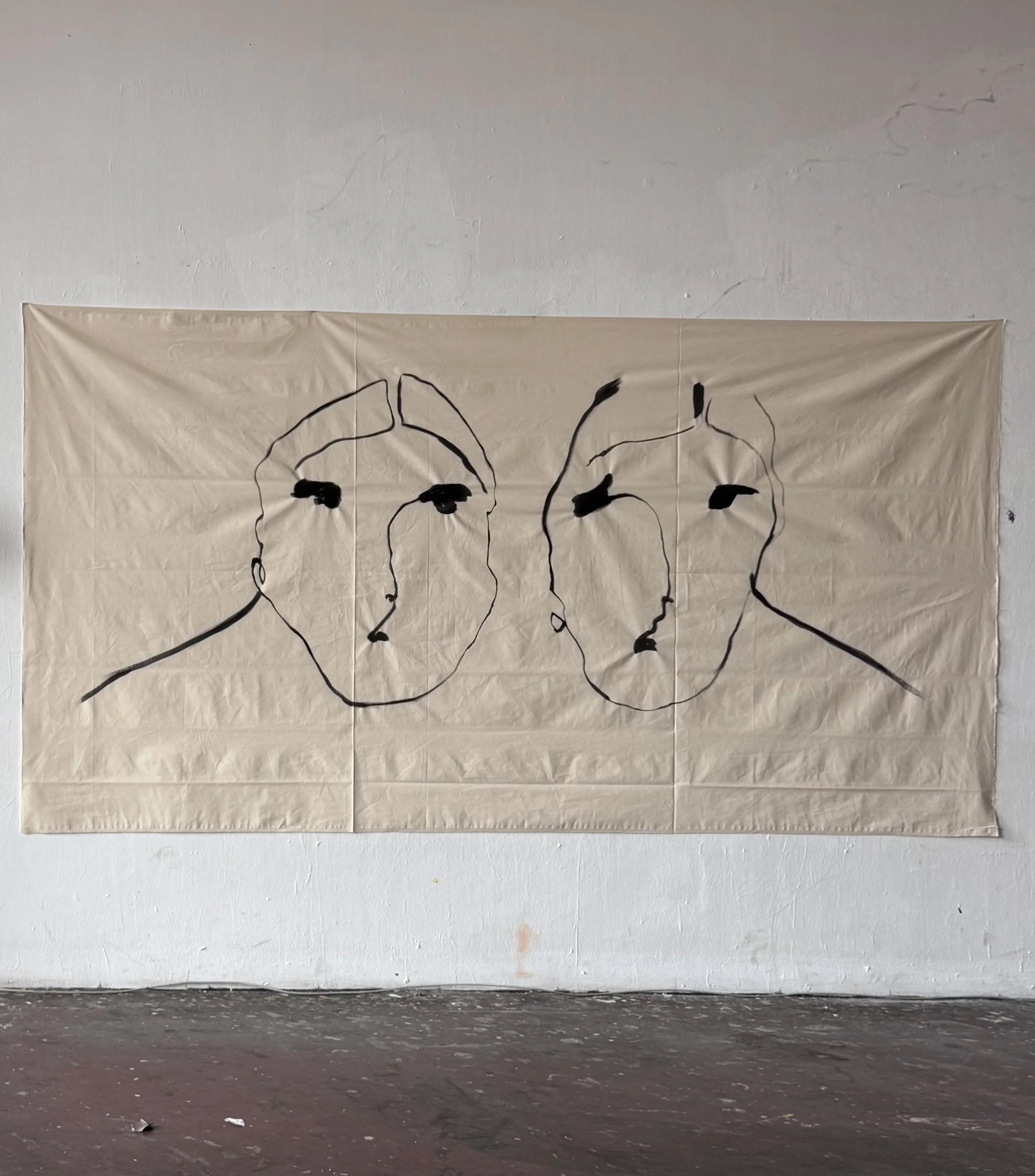 A large beige cloth with a minimalistic black line drawing of three faces attached to a white wall. The faces are outlined with simple black lines with some features shaded in black. The wall has some textured imperfections and slight cracks.