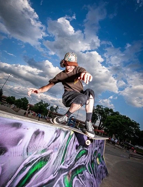 Young skateboarder performing a trick on a graffiti-covered ramp outdoors under a partly cloudy sky.