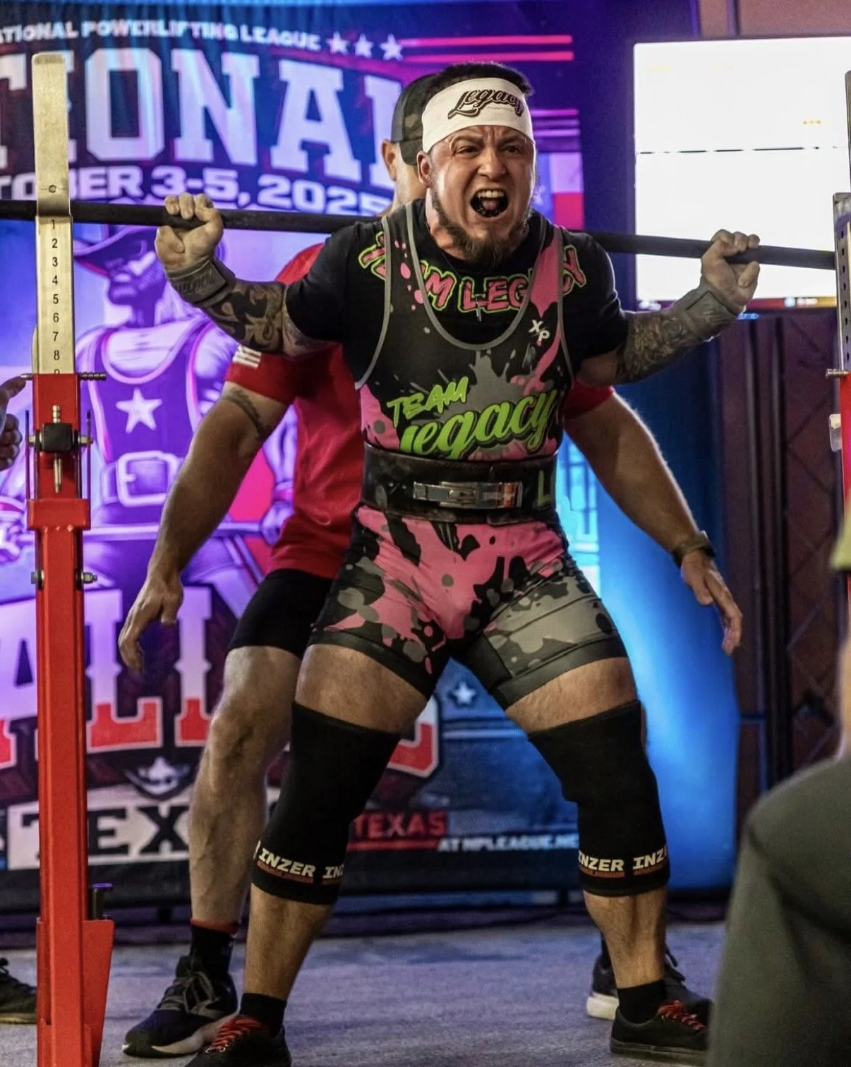 A man participating in a powerlifting competition, performing a squat. He is wearing a colorful effortment with 'Team Legacy' printed on it, a white headband with 'Legacy' written on it, and black knee-high socks with the brand 'Inzer'. He appears strained as he lifts a barbell loaded with weights, with a competitive backdrop and spectators nearby.