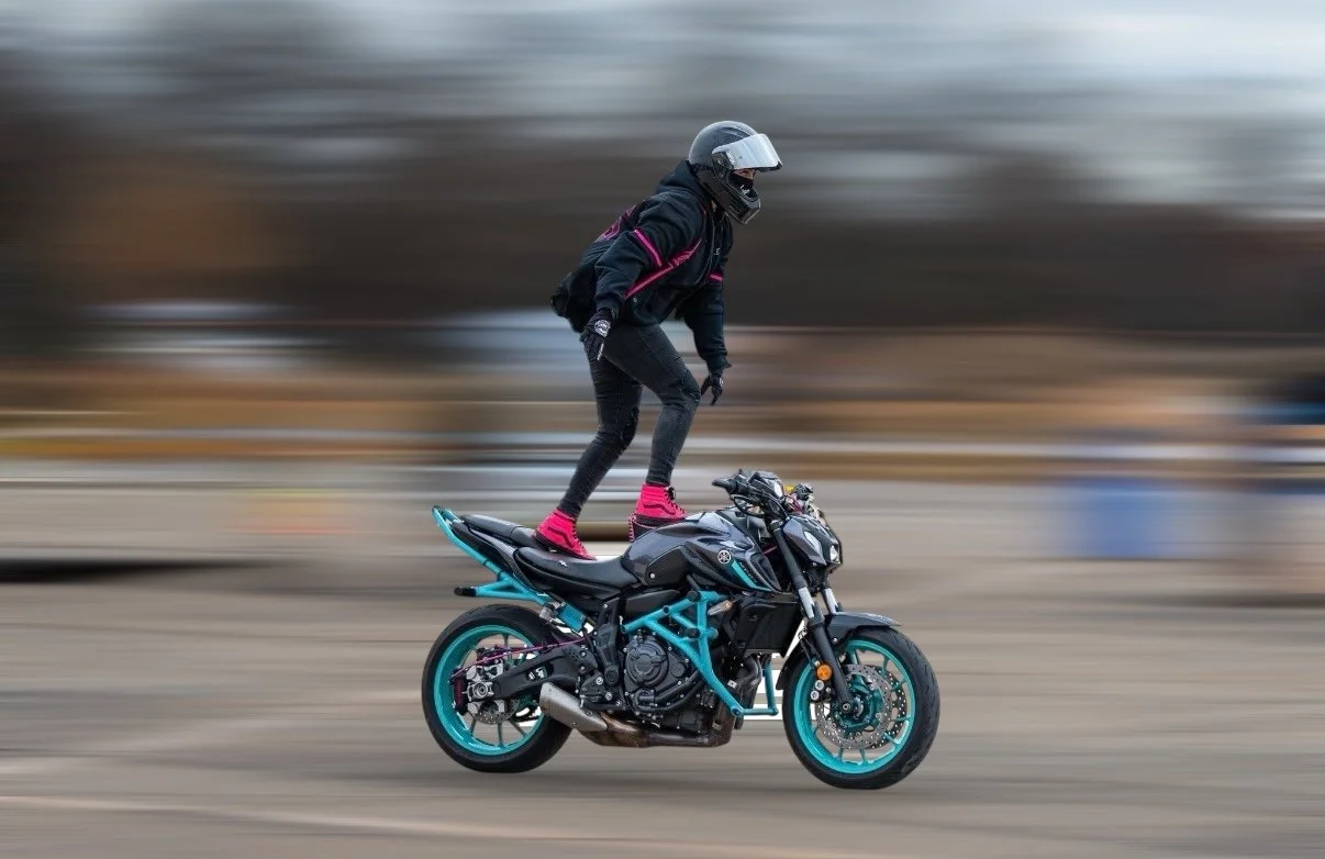 Person in black riding gear and helmet balancing on a motorcycle while skateboarding.