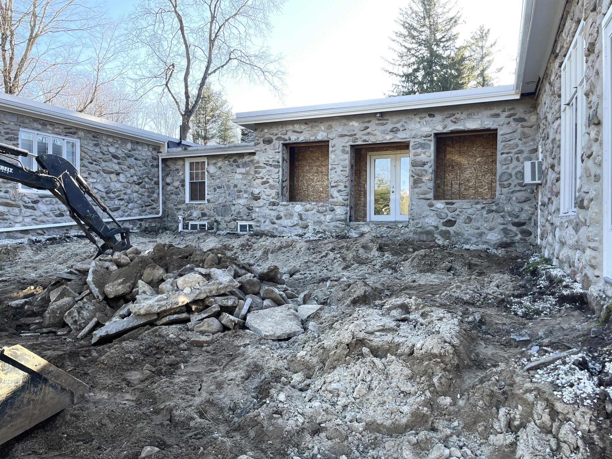 Exterior home renovation in progress showing demolition of back wall, removal of slate patio, and disturbed landscaping before new outdoor living space installation.