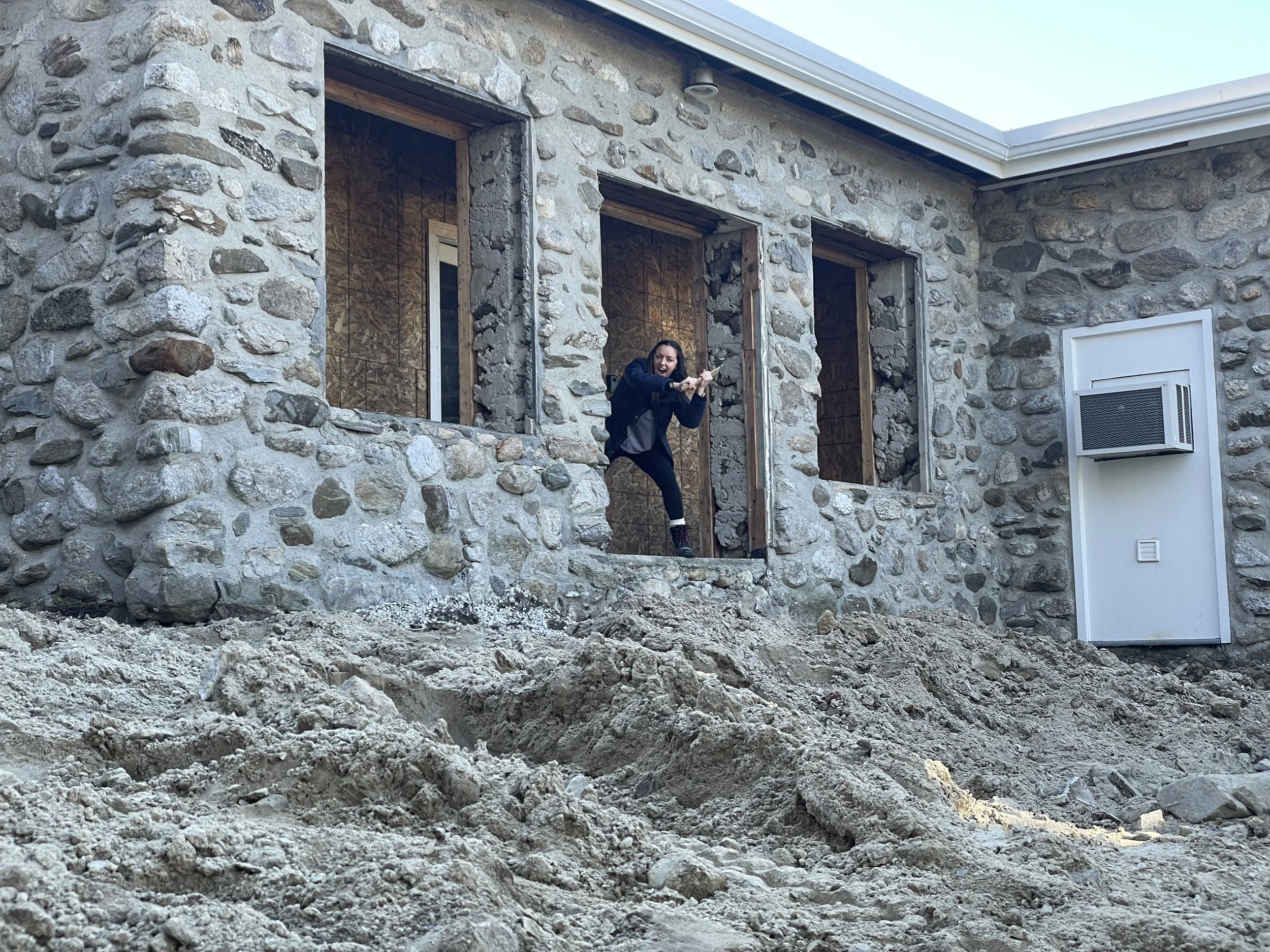 Exterior home renovation in progress showing demolition of back wall, removal of slate patio, and disturbed landscaping before new outdoor living space installation.