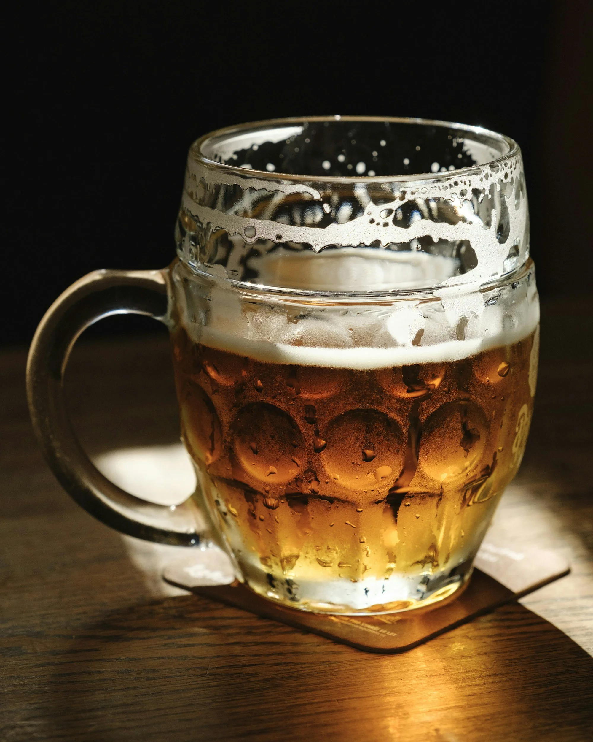 A glass mug filled with amber beer on a wooden surface, with foam remnants on the inside of the glass and condensation on the outside.
