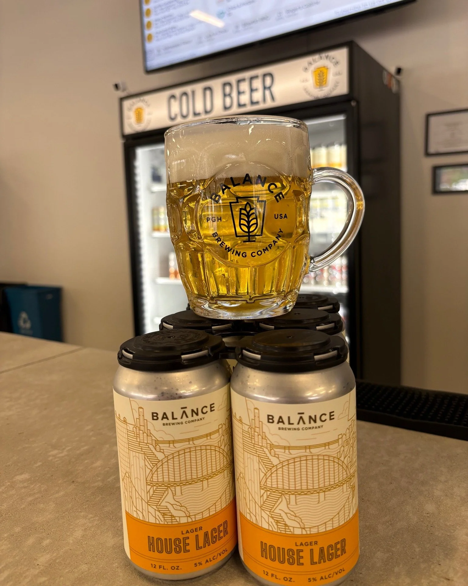 A 6 pack of yellow and white cans with a line drawing of a Pittsburgh bridge on it. A handled mug full of a light orange beer is balanced on top. A large beer cooler is visible in the background. 