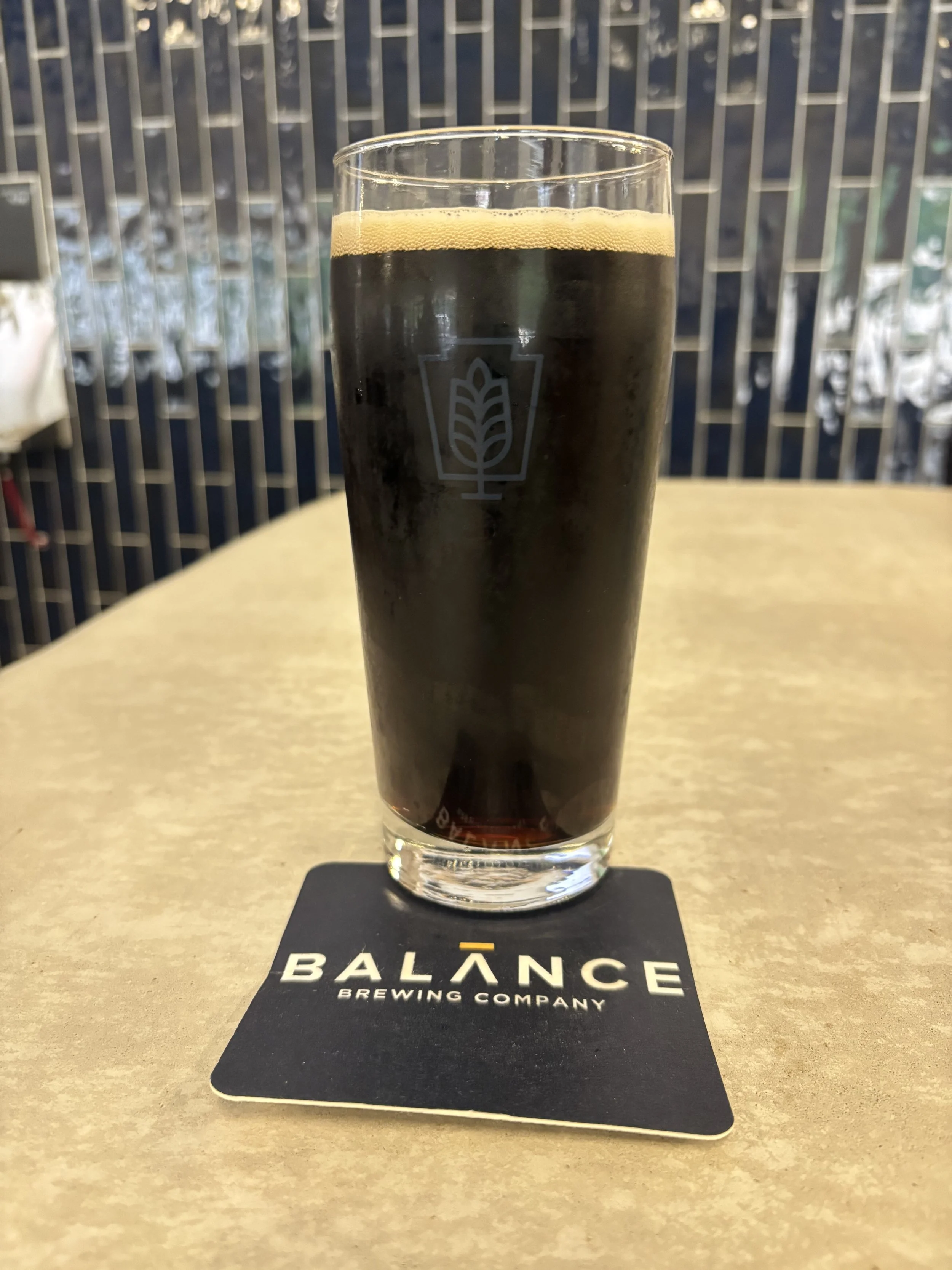 A photograph of full glass of a dark brown beer in front of a shiny blue tile wall. The coaster beneath displays the Balance Brewing logo.
