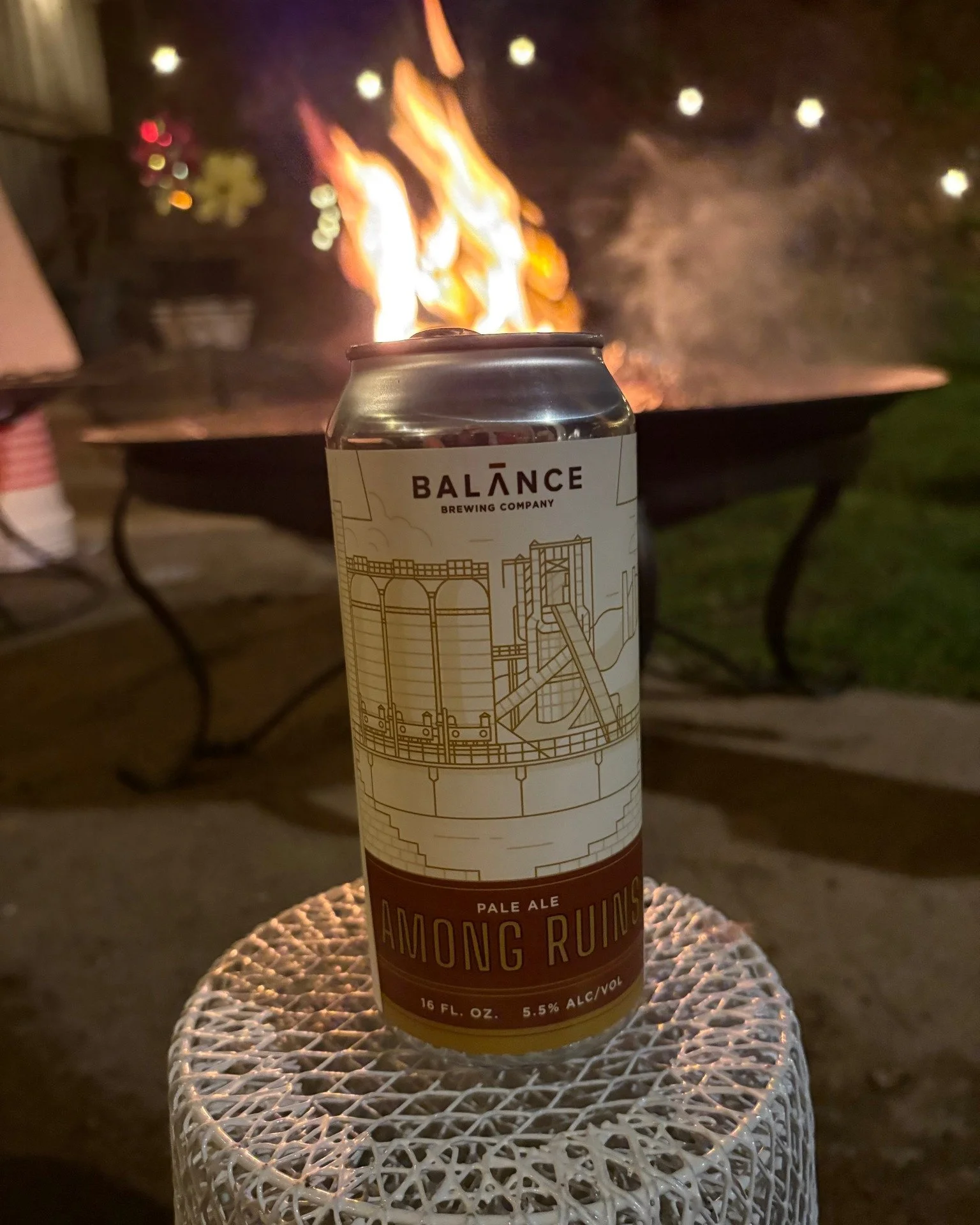 A tan and brown can with line drawings of a factory sits on a table; a firepit in the background makes it look like flames are coming out of the top of the can. Very dramatic. 