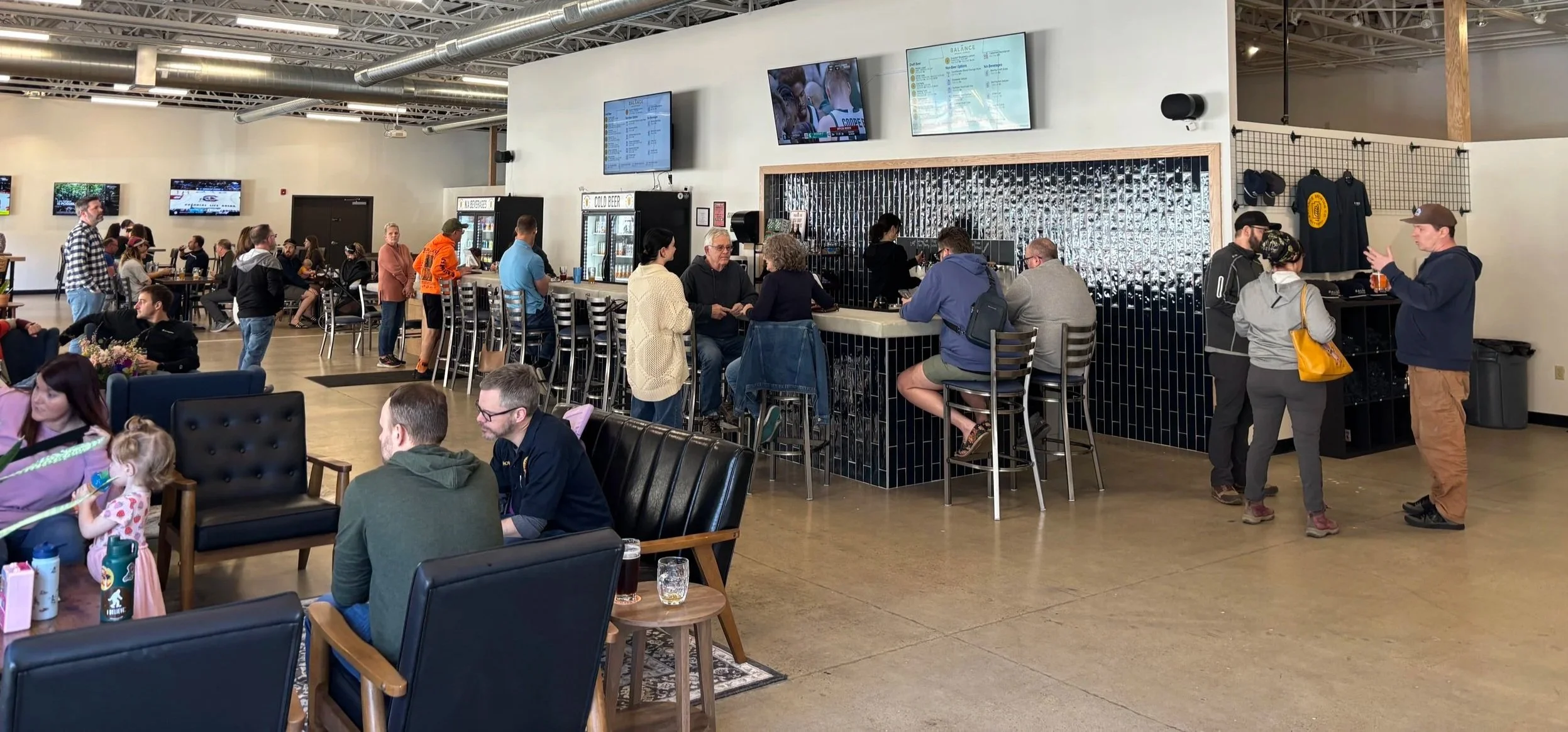 Photograph of a busy tap room, with customers standing, sitting at the bar and sitting in couches.