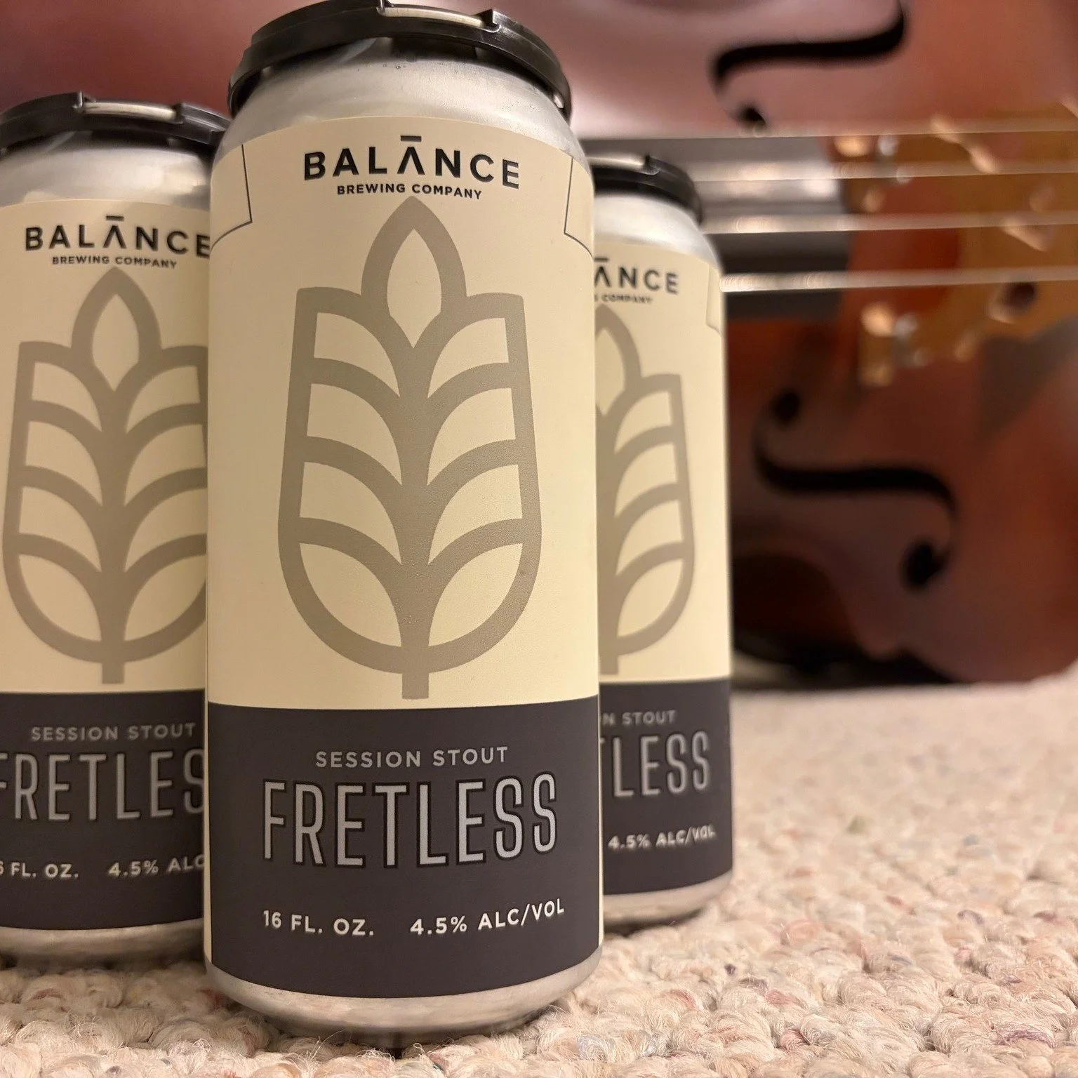 A 4 pack of Fretless cans, a white can with black markings, sits on a carpeted floor; a violin lays on its side in the background. 