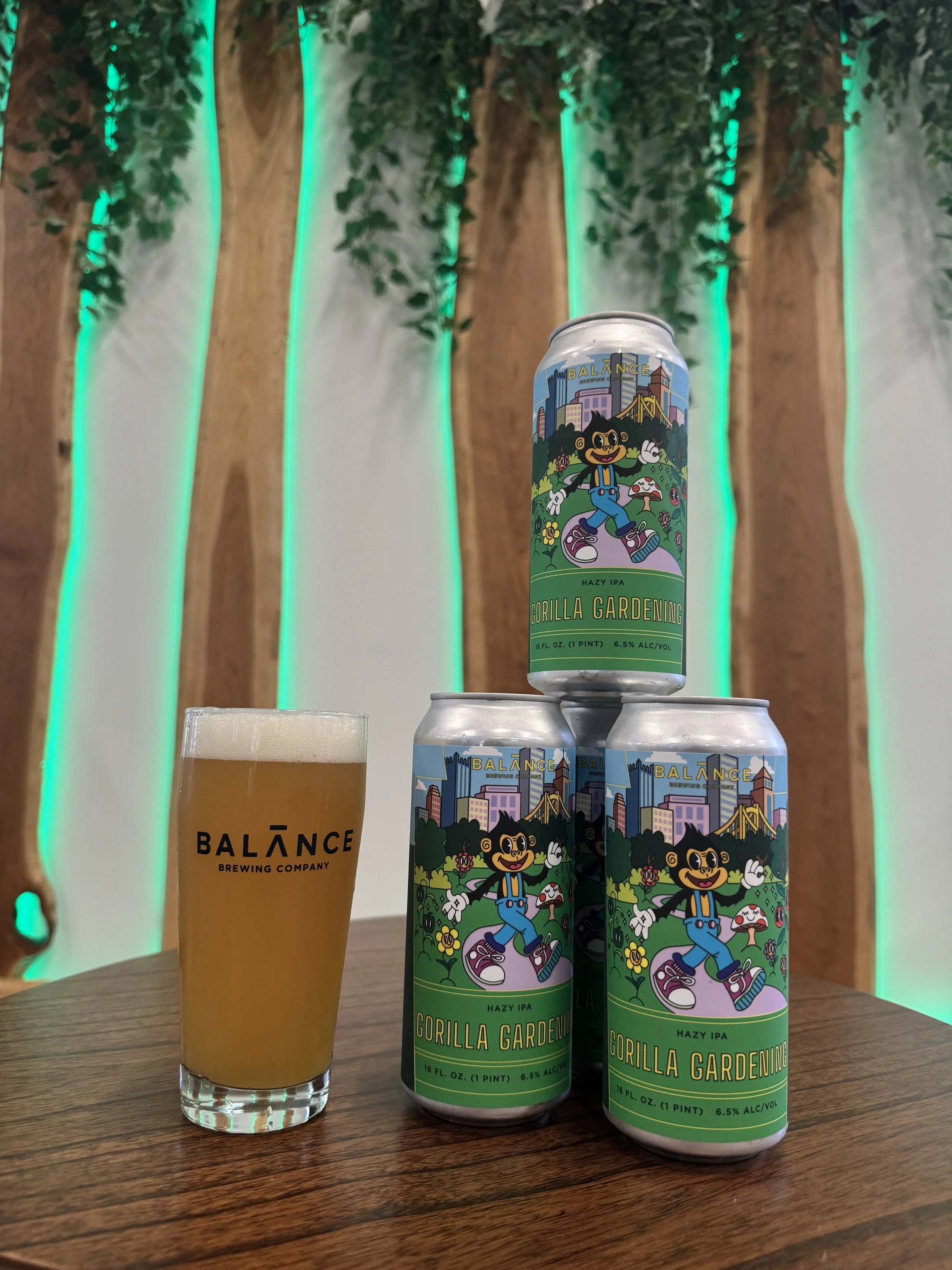 Orange beer in a glass, next to 3 cans stacked on top of each other, in front of green lit wood panels.