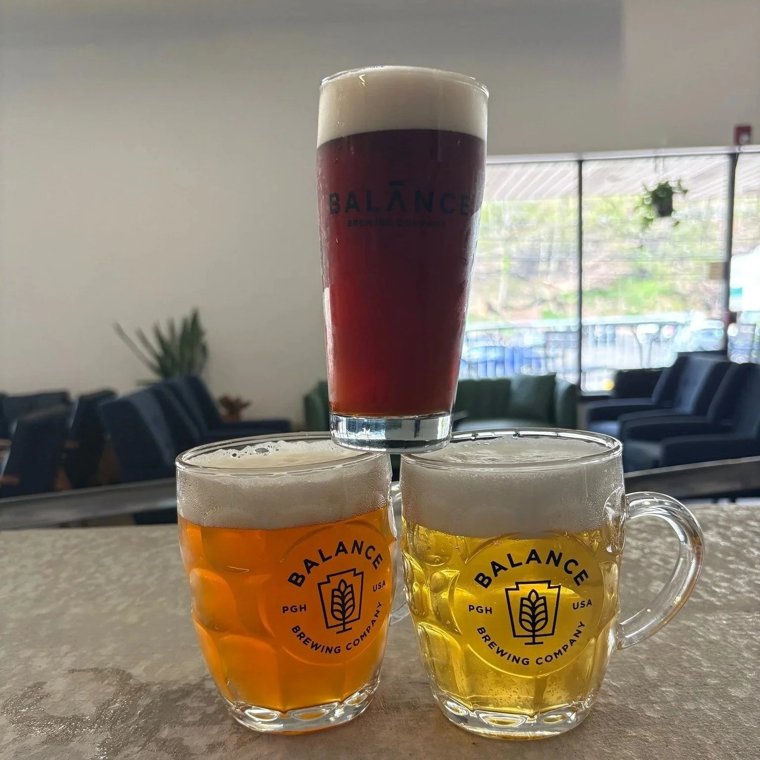 Photograph of a tower of beers. Two mugs of beer, one lighter  yellow, one a medium orange sit on a gray bar. Balanced where the rims of the mugs meet is a darker brown ale. All three of the glasses have the Balance Brewing logo.