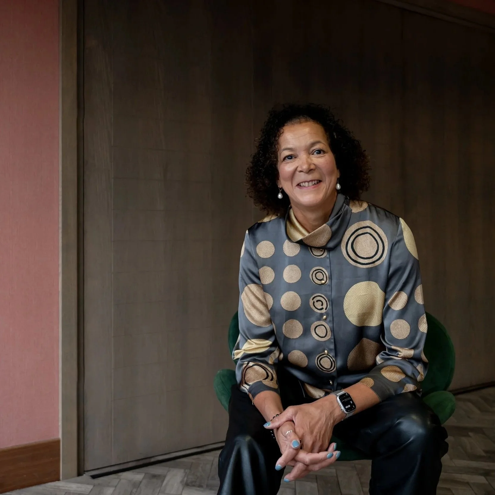 A woman with curly dark hair smiling while sitting on a green chair in front of a dark wooden wall. She is wearing a shiny blouse with a circular pattern in gold, gray, and black, and a smartwatch on her left wrist.