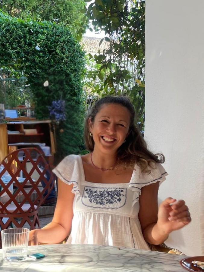 A woman with long brown hair smiling and sitting at a marble table outdoors in a garden setting.