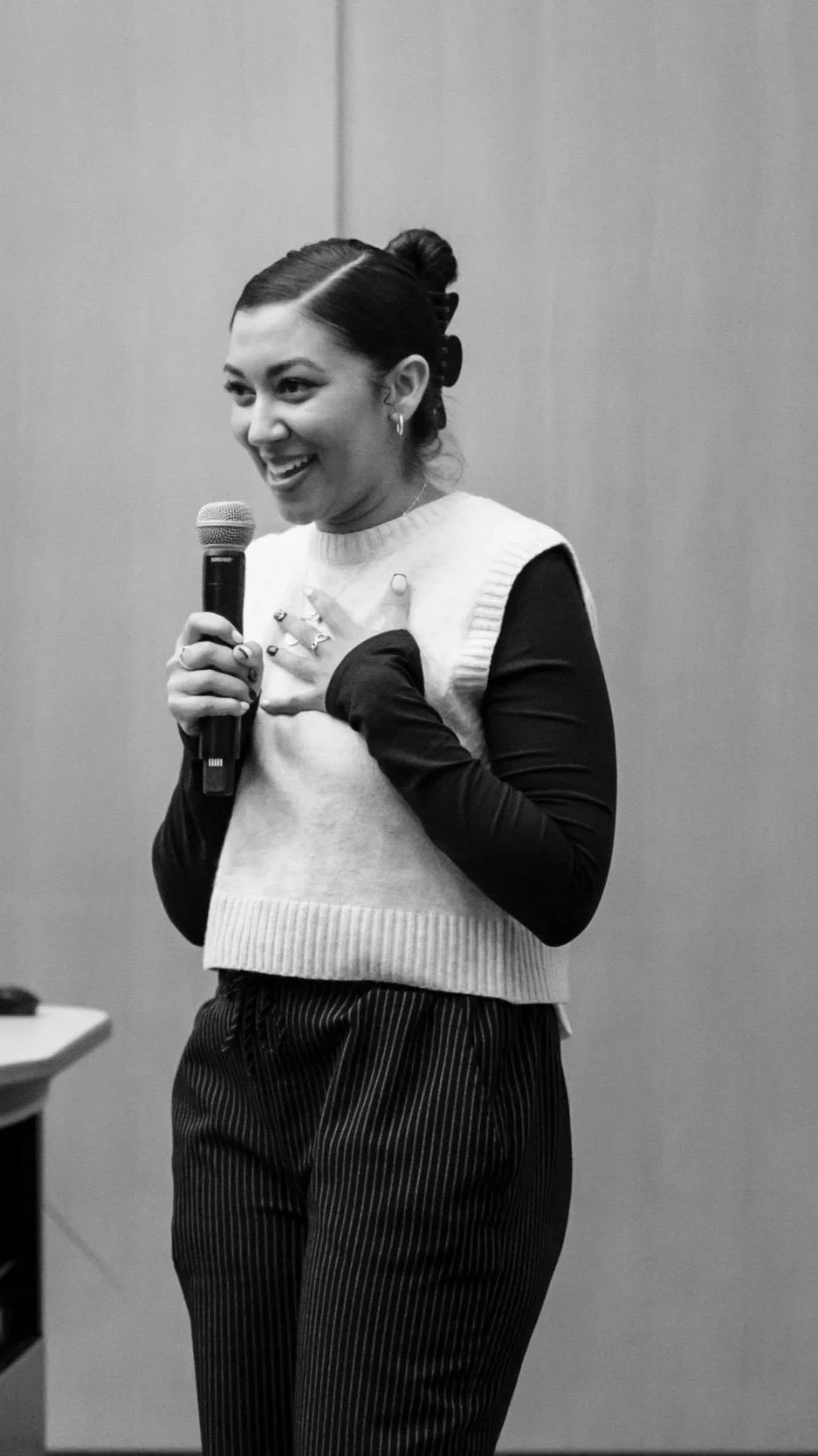 Black and white photo of a smiling woman speaking into a microphone, wearing a sleeveless sweater with long black gloves, and gesturing with one hand on her chest.