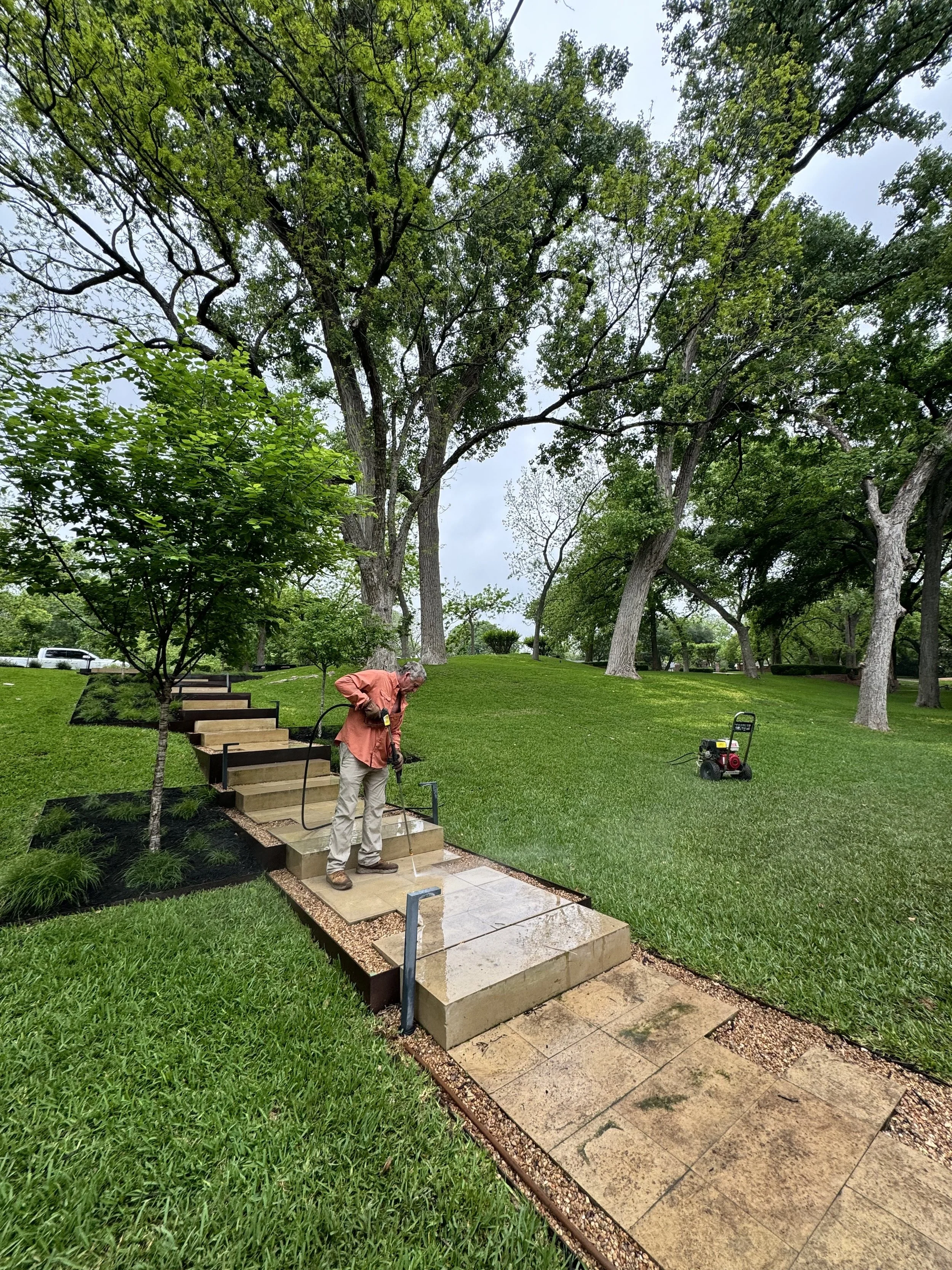 A man watering newly installed outdoor stone steps in a lush green park with tall trees and a cloudy sky.