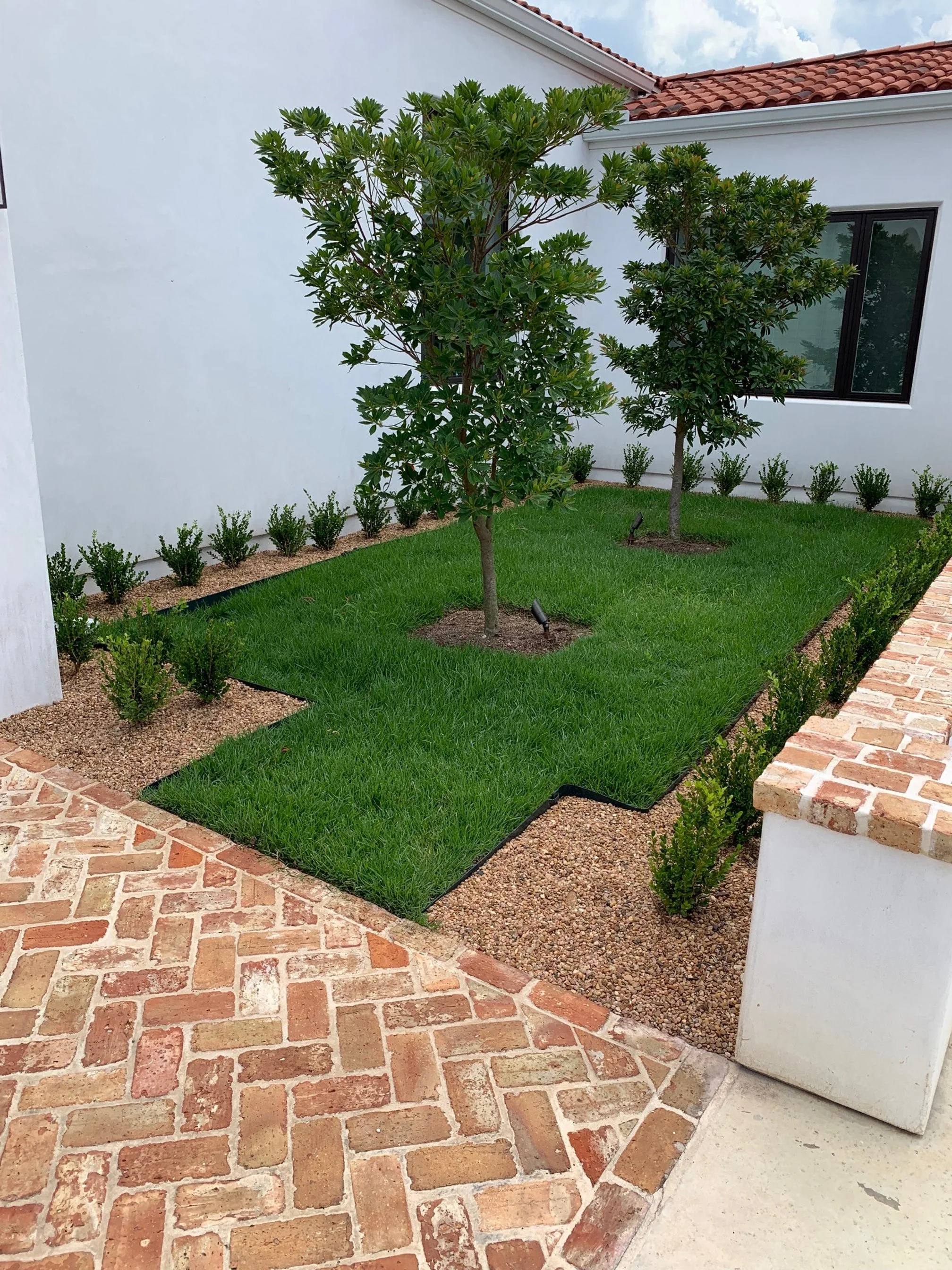 A landscaped backyard with a brick-paved patio, green grass, small bushes, two large trees, and white exterior walls with a window.