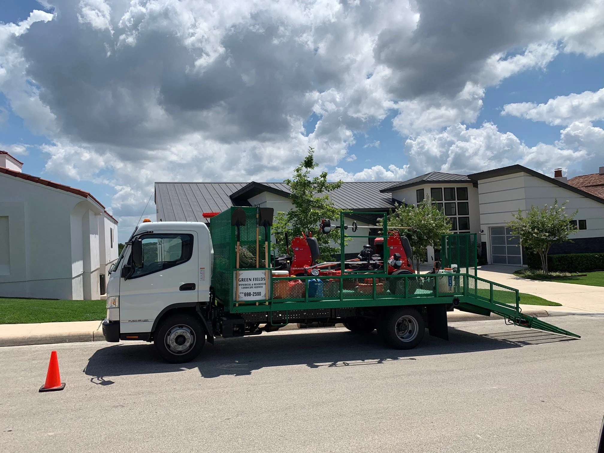 A white flatbed truck with garden and landscaping equipment on the back, parked on a suburban street under partly cloudy sky with houses and trees in the background.