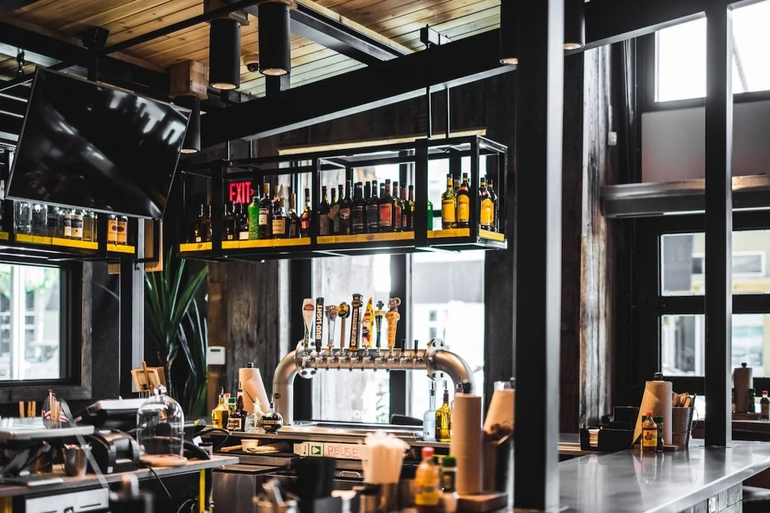Interior of a modern bar with a black metal and wooden decor, bottles of alcohol on shelves, a beer tap, and a flat-screen TV mounted on the wall.