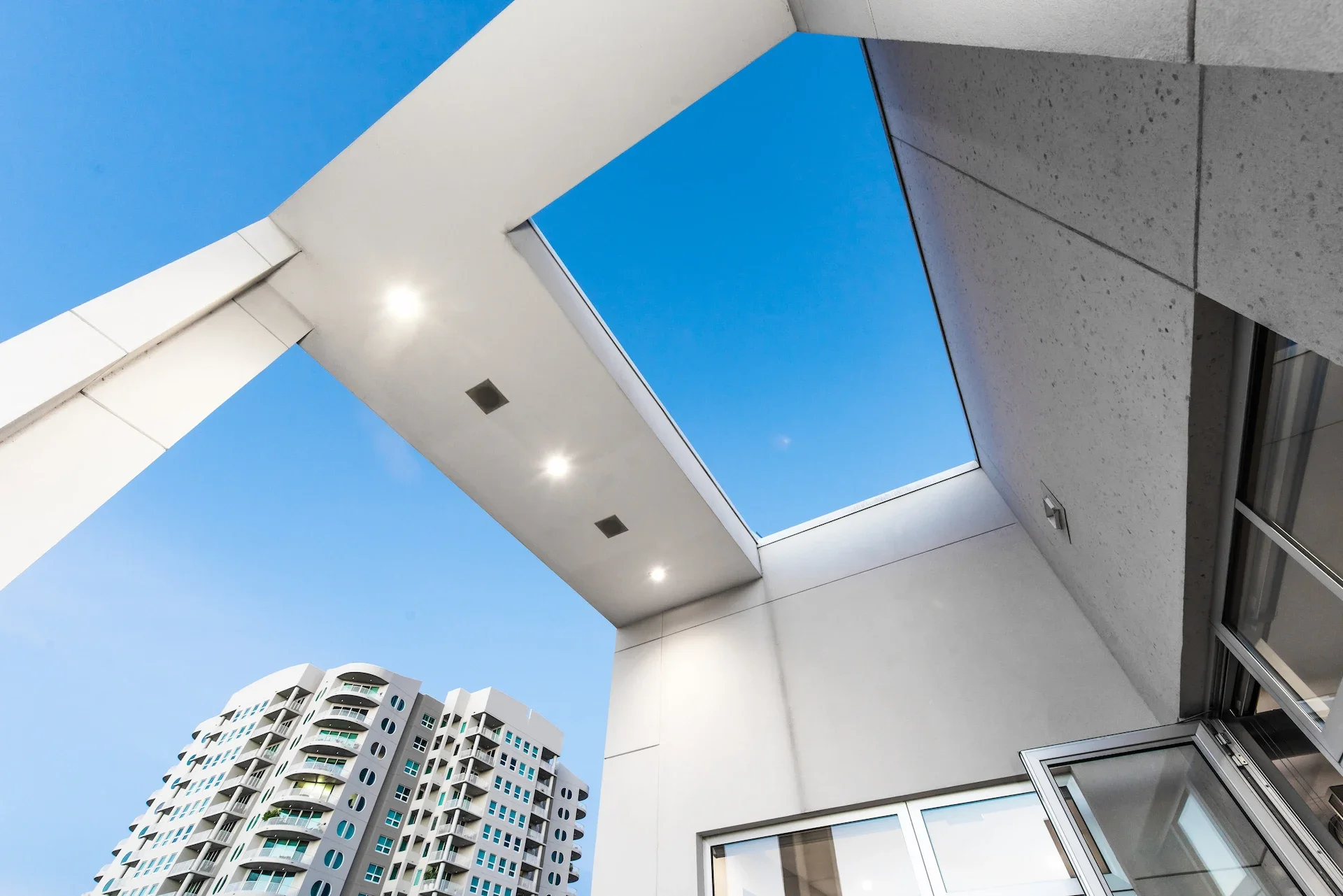 Looking up at a modern building with a square roof opening, exposing a clear blue sky, and a in the background, a high-rise residential building with curved balconies.