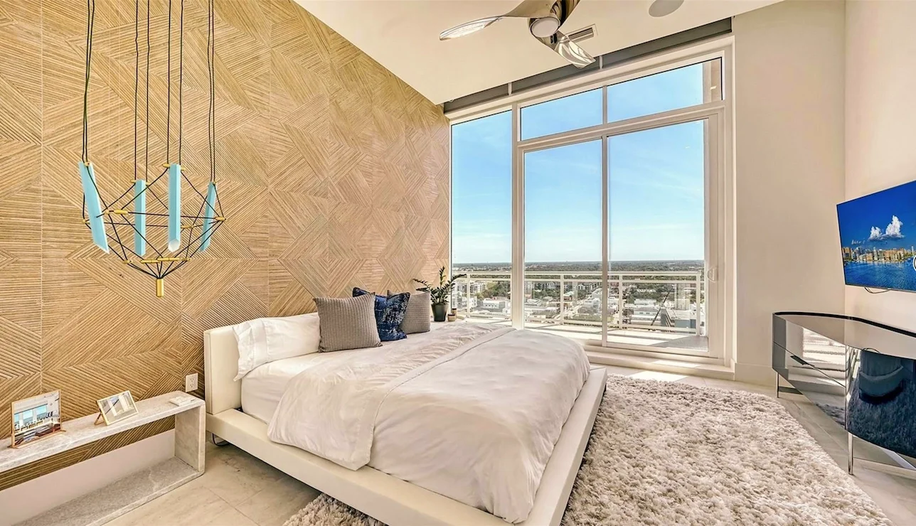 Modern bedroom with large window view, white bed with gray and blue pillows, wooden accent wall, beige shaggy rug, TV, and decorative chandelier.