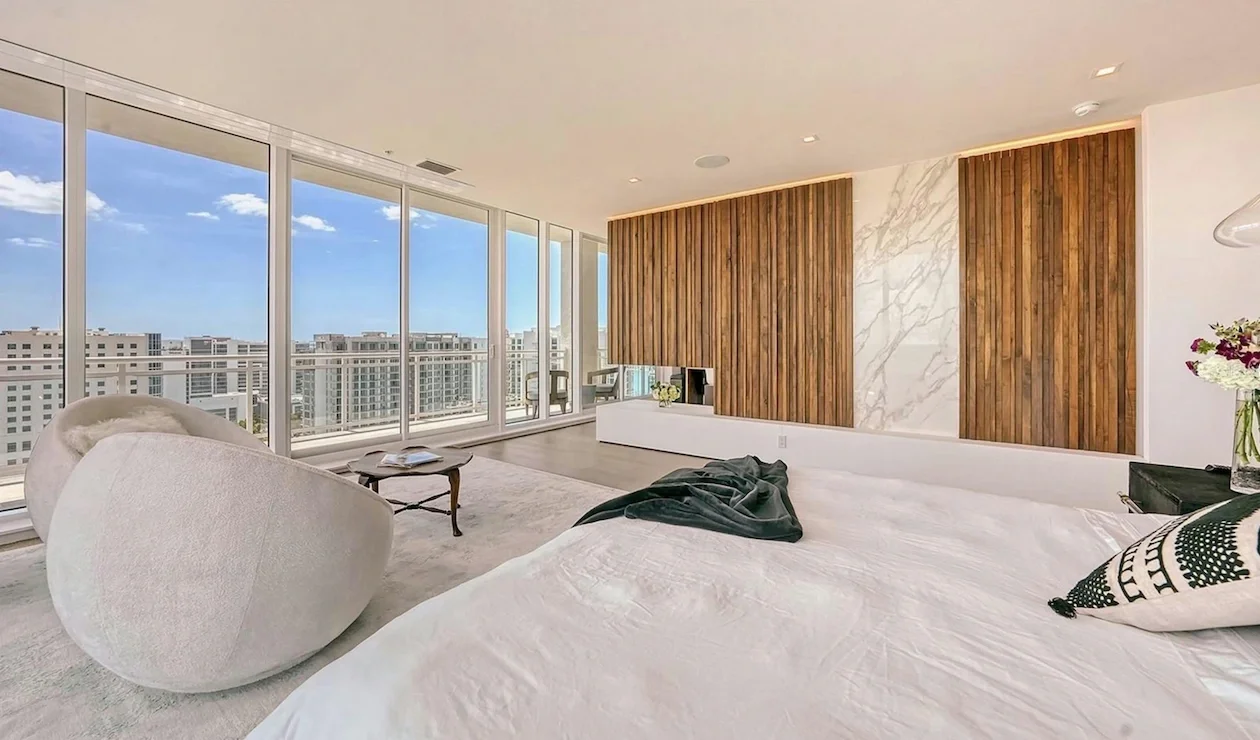A modern bedroom with large floor-to-ceiling windows showing a cityscape with tall buildings and a blue sky. The room features a white bed with a black throw blanket and decorative pillows, a small round table with books, a chair, and wooden and marble wall panels.