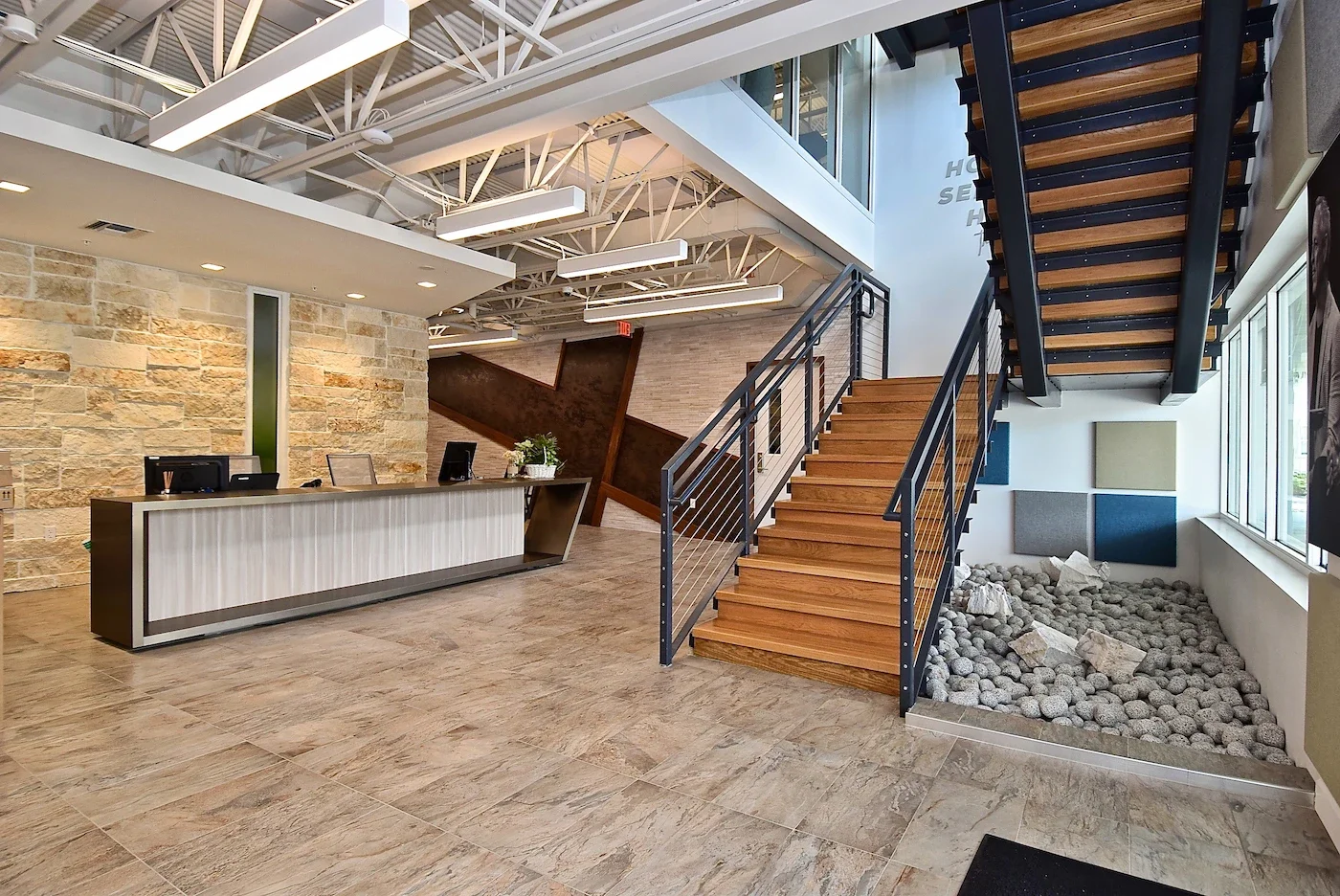 Modern office reception area with stone wall, wooden staircase, and large windows, featuring a check-in desk with computers and a decorative pebble bed with wall art.