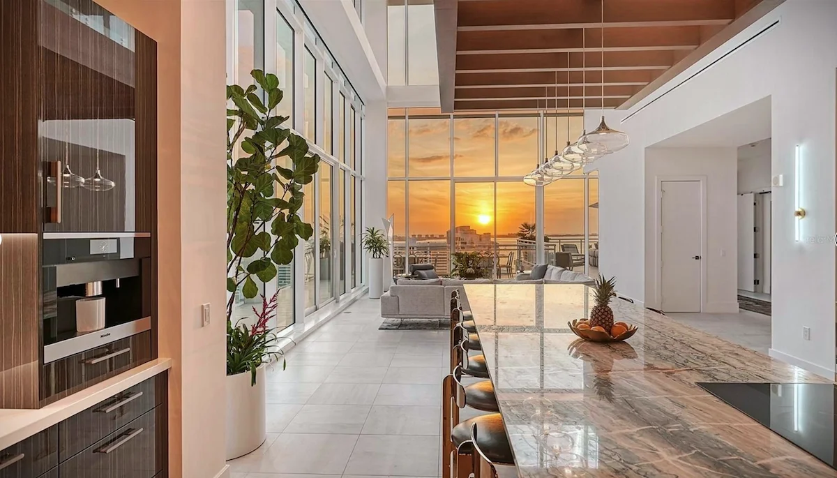 Modern apartment interior with a large marble kitchen island, high ceilings, large windows with a sunset view, and a seating area with couches and plants.