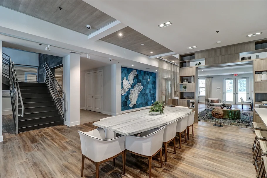 Modern open-concept interior with a marble dining table surrounded by white chairs, a wooden floor, a staircase with metal railing, a blue textured wall with large white fish decor, and a sitting area with chairs and a rug in the background.
