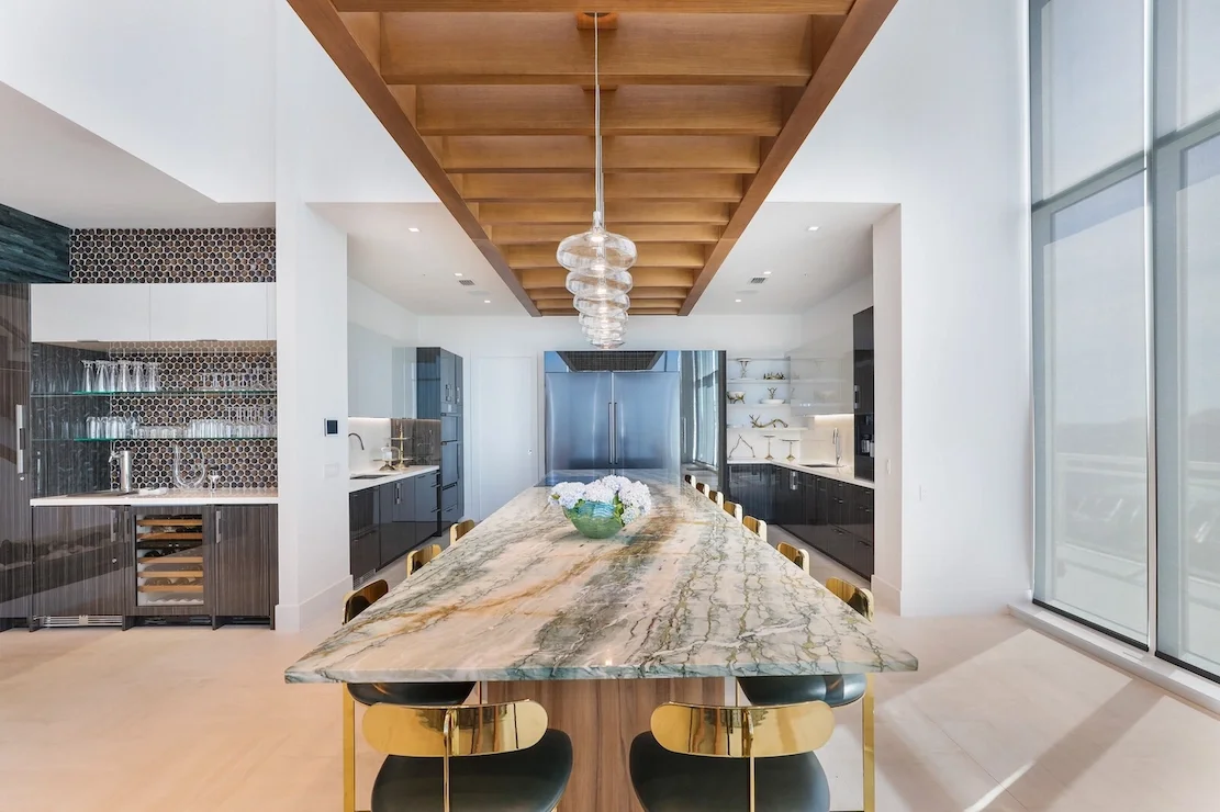 Modern kitchen with a large marble dining table, black and gold chairs, wooden ceiling, and glass pendant lighting.