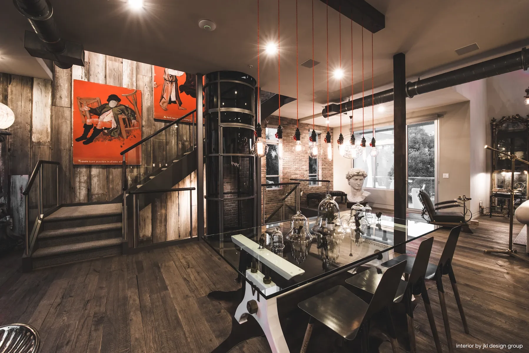 Interior of a contemporary living space with wooden flooring, brick and wood wall decor, modern lighting fixtures hanging from the ceiling, art posters on the walls, a glass dining table with decorative glass cloches, and a classical bust sculpture near large windows.