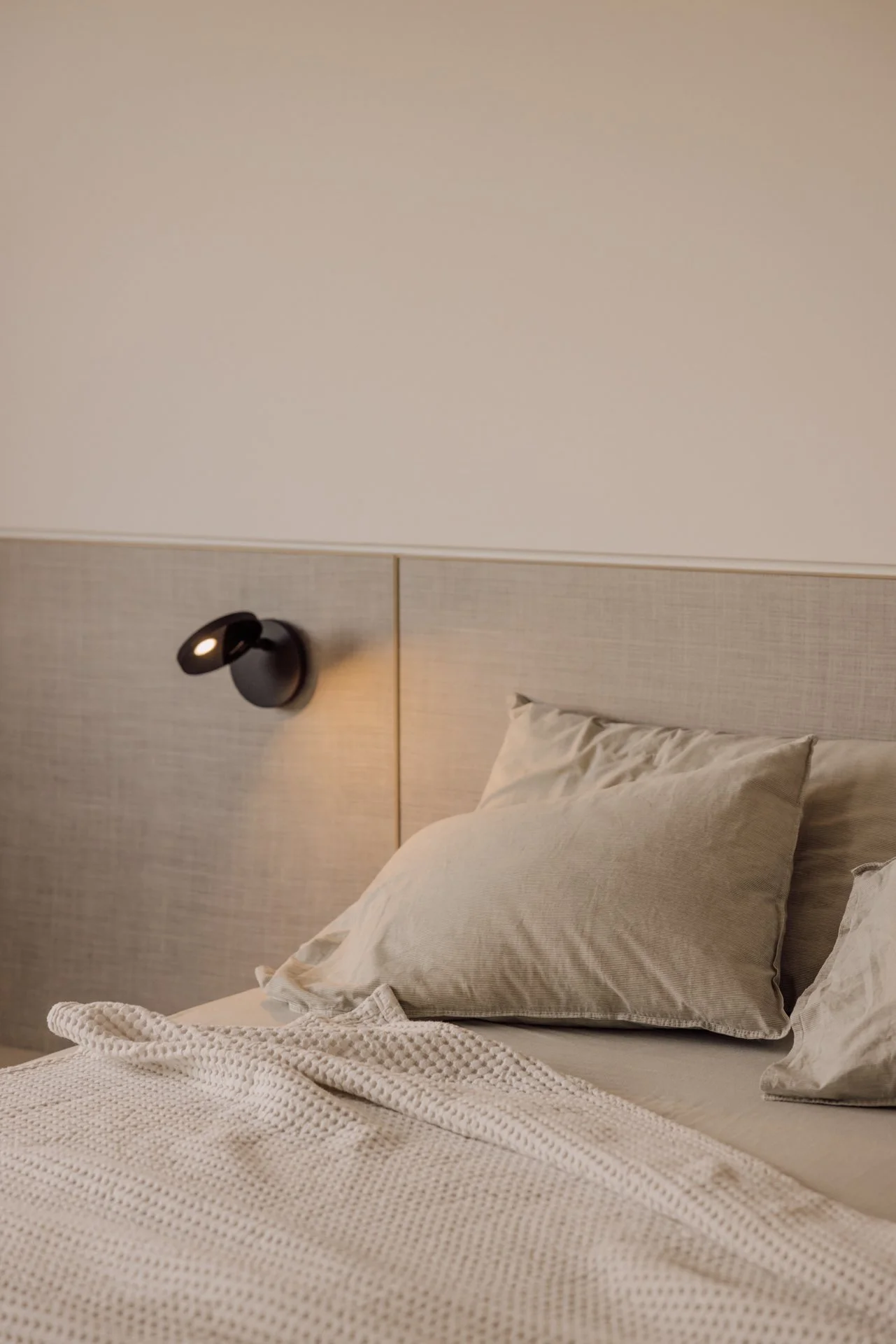 Close-up of a neatly made bed with beige pillows, set against a neutral-tone headboard and wall, with a black adjustable reading light attached to the wall.