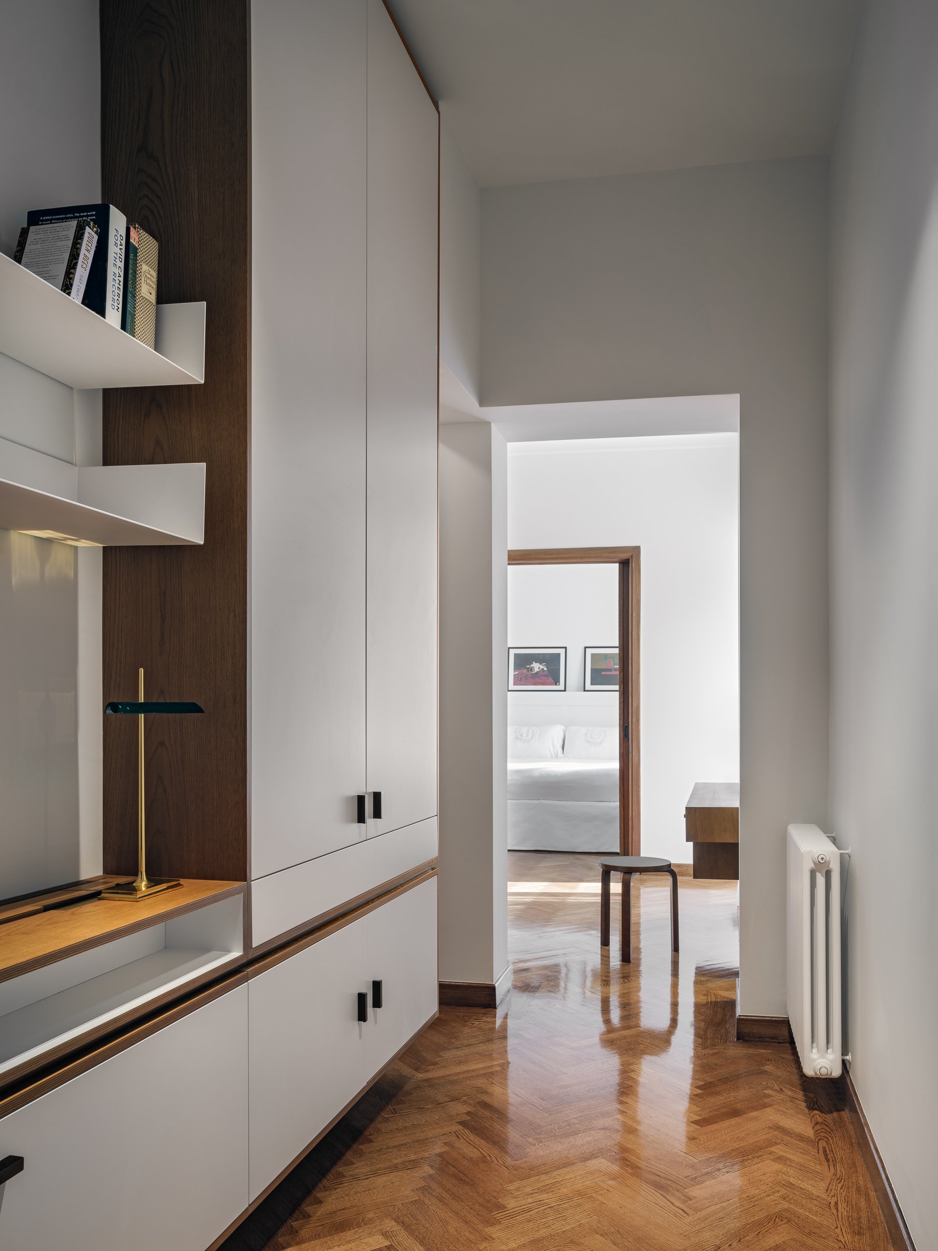 A hallway with wooden flooring and white walls leading to a living room with a white sofa and framed artwork. There are built-in cabinets and shelves on the left and a radiator on the right side.