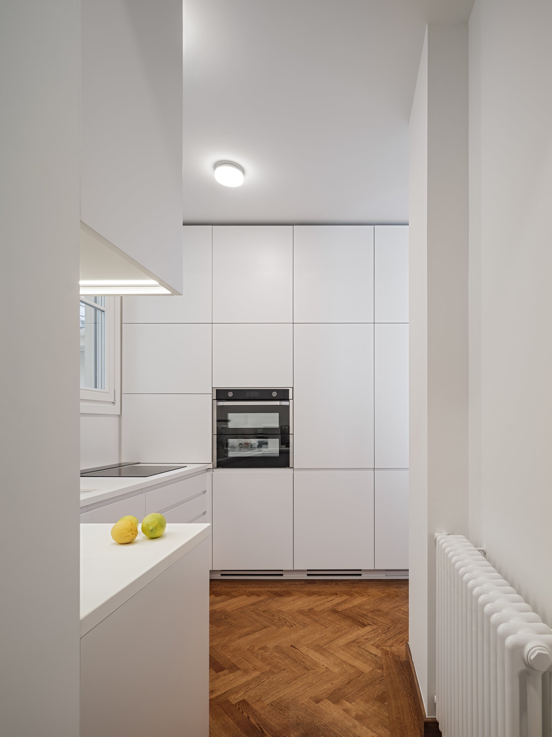 Minimalist white kitchen with built-in oven, white cabinets, a window, and a wooden floor. Two fruits are on a white countertop.