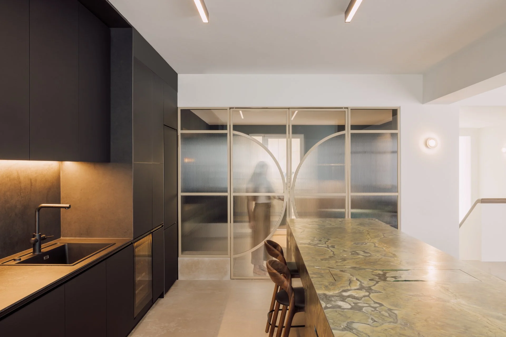Modern kitchen with black cabinetry, beige countertop, and a backsplash with warm lighting. There is a marble or stone dining table with wooden chairs. A glass partition with circular design separates the kitchen from another room.