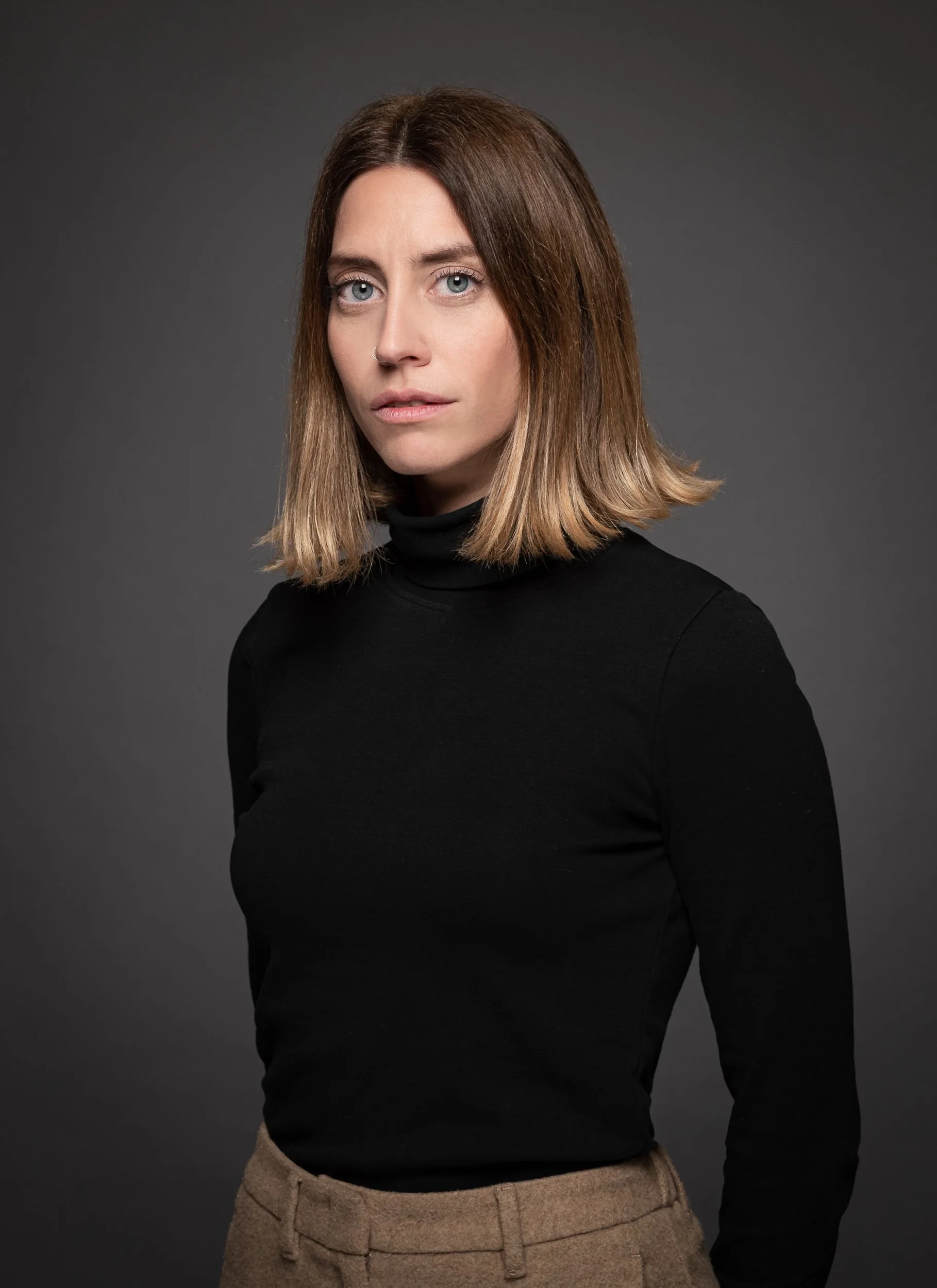 Portrait of a woman with shoulder-length light brown hair, wearing a black turtleneck and beige trousers, standing against a dark gray background.