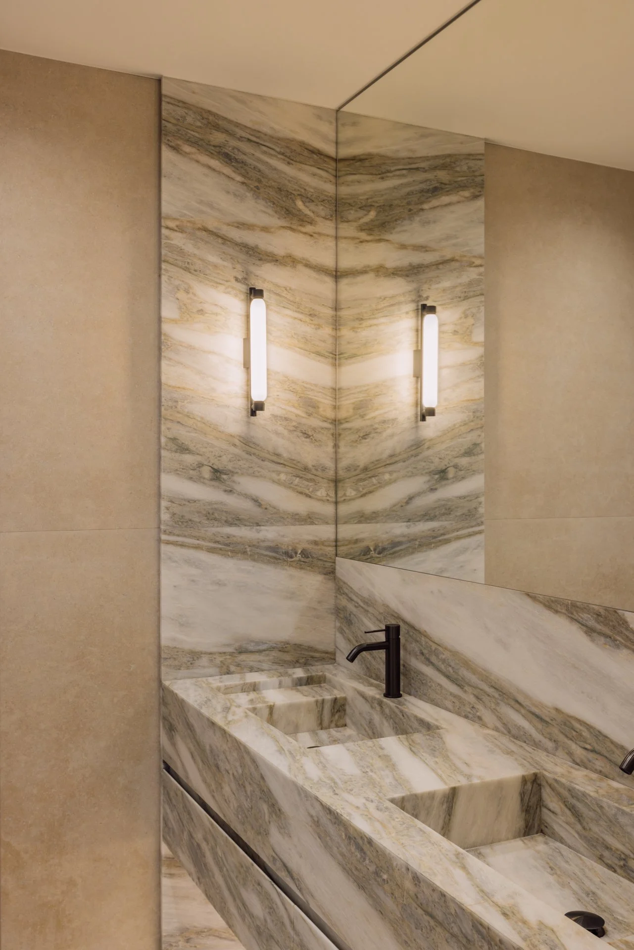 A modern bathroom with a marble double sink vanity, beige walls, and two vertical wall-mounted light fixtures.