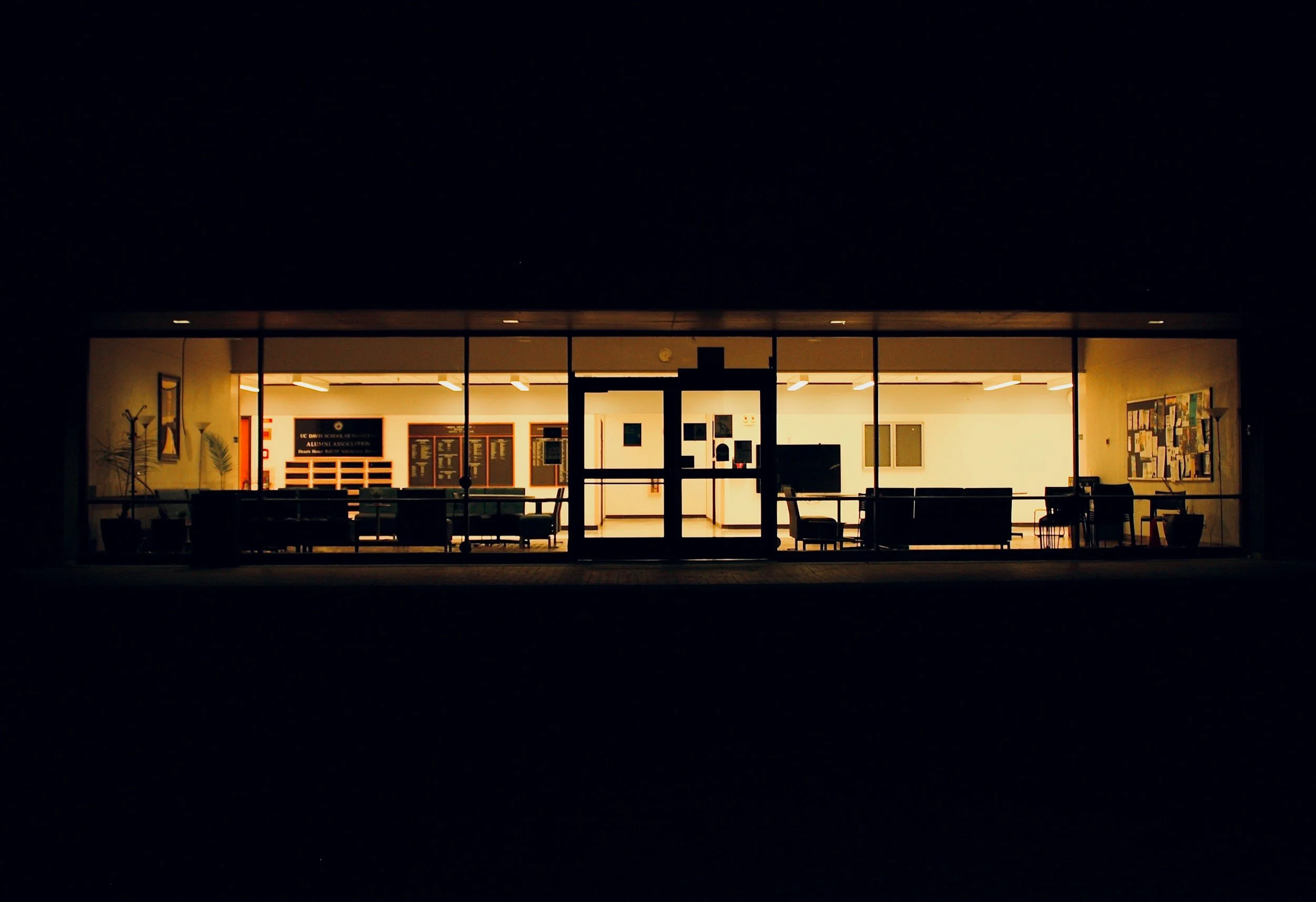 Illuminated interior of a school or community center seen through large window at night, with seating and bulletin boards inside.