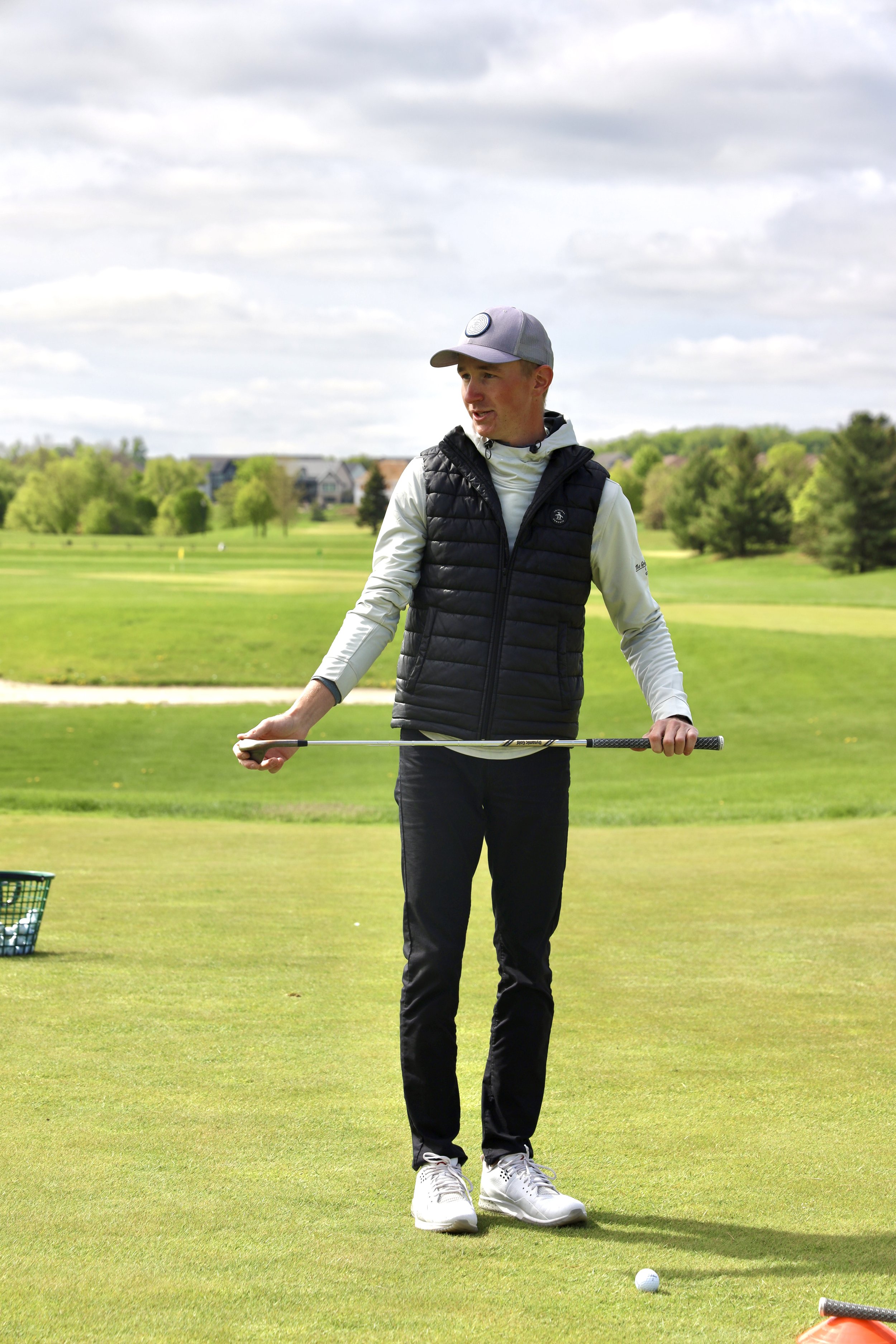 A young man in a golf course holding a golf club, dressed in a gray cap, white long sleeve shirt, black quilted vest, black pants, and white shoes, preparing to hit a golf ball.