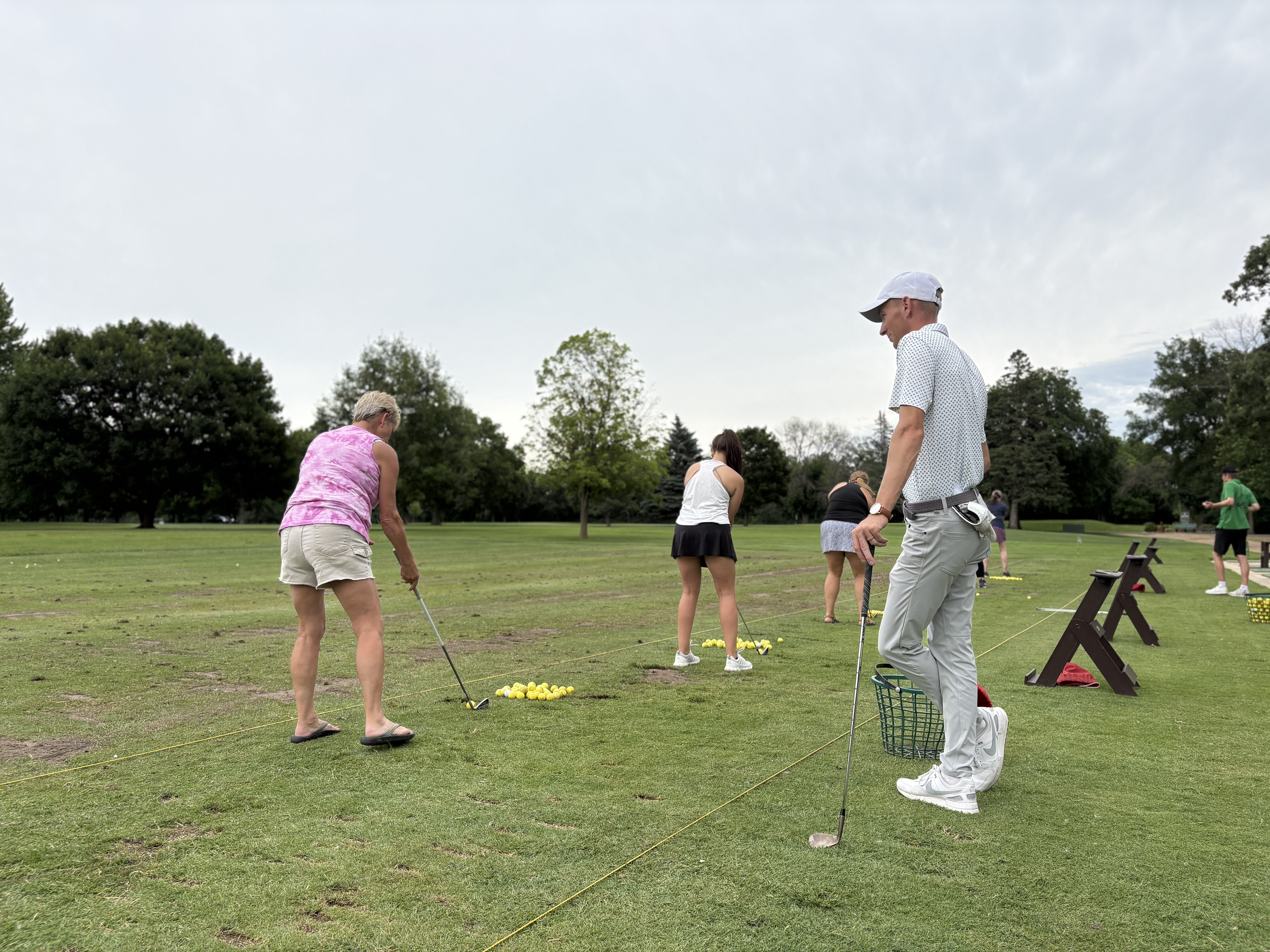 People practicing golf on a driving range with yellow golf balls, grassy field, trees, and overcast sky in the background.