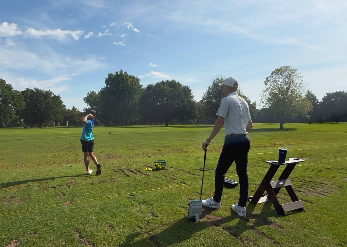 A man and a woman practicing golf on a grassy field with trees in the background under a clear blue sky. The man is standing near a small table with a drink, while the woman is swinging a golf club.