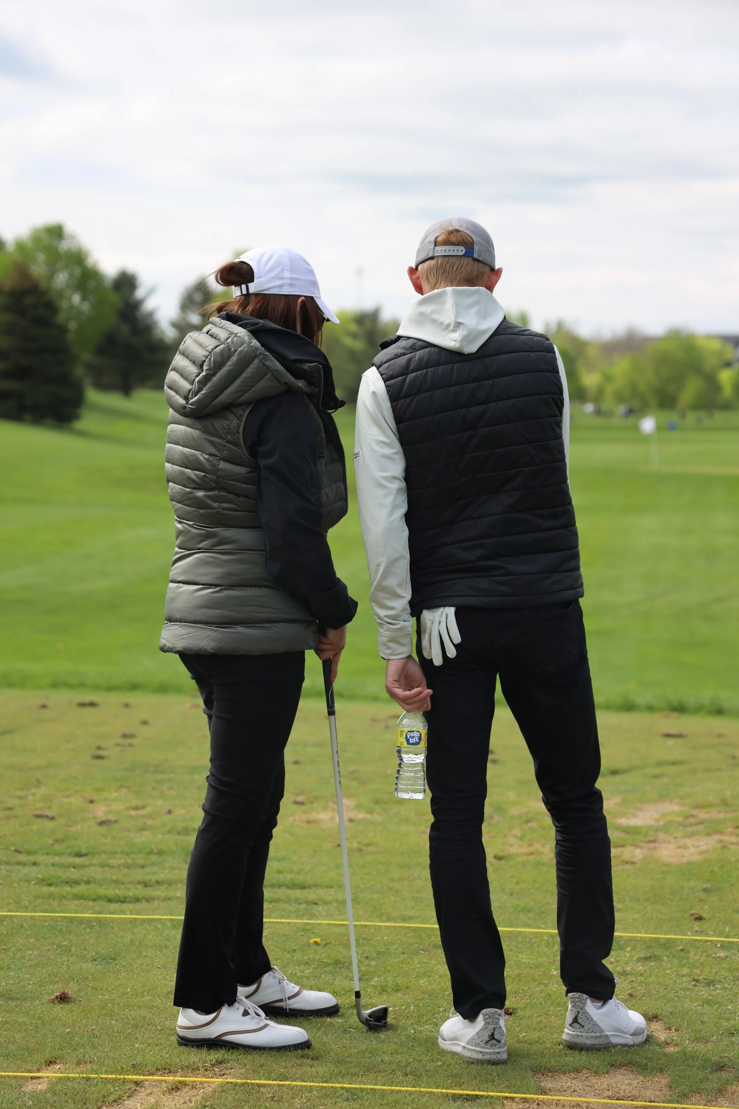 Two golfers, a woman and a man, standing on a golf course discussing their game. The woman is holding a golf club and is wearing a white cap, black and grey jacket, black pants, and white golf shoes. The man is holding a water bottle, wearing a gray cap backwards, black vest over white hoodie, black pants, and white athletic shoes. They are standing on the grass near yellow markers with trees and a partly cloudy sky in the background.