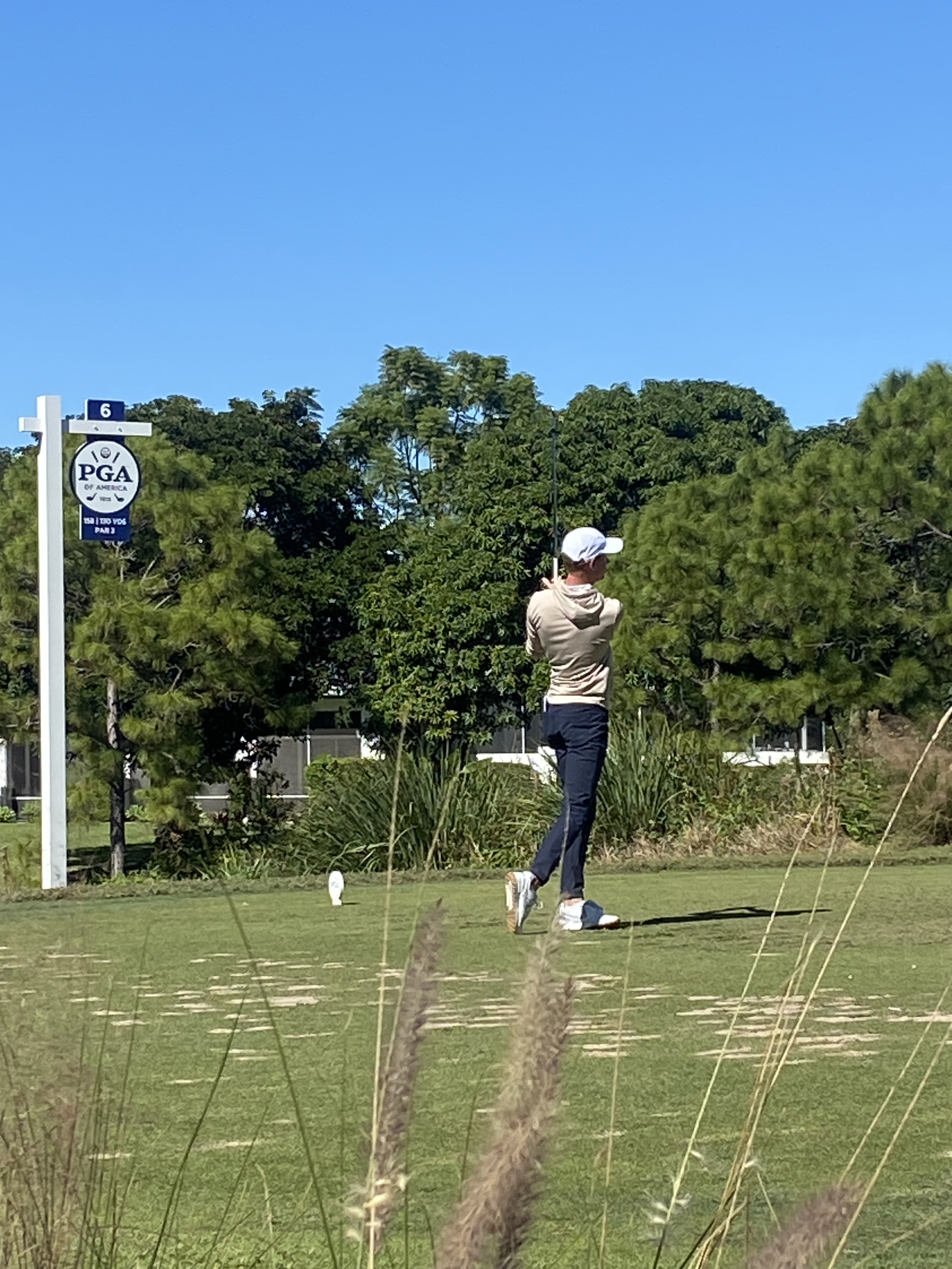 A person in a white cap and beige hoodie taking a shot on a golf course under a clear blue sky, with trees and a sign for the PGA of America in the background.