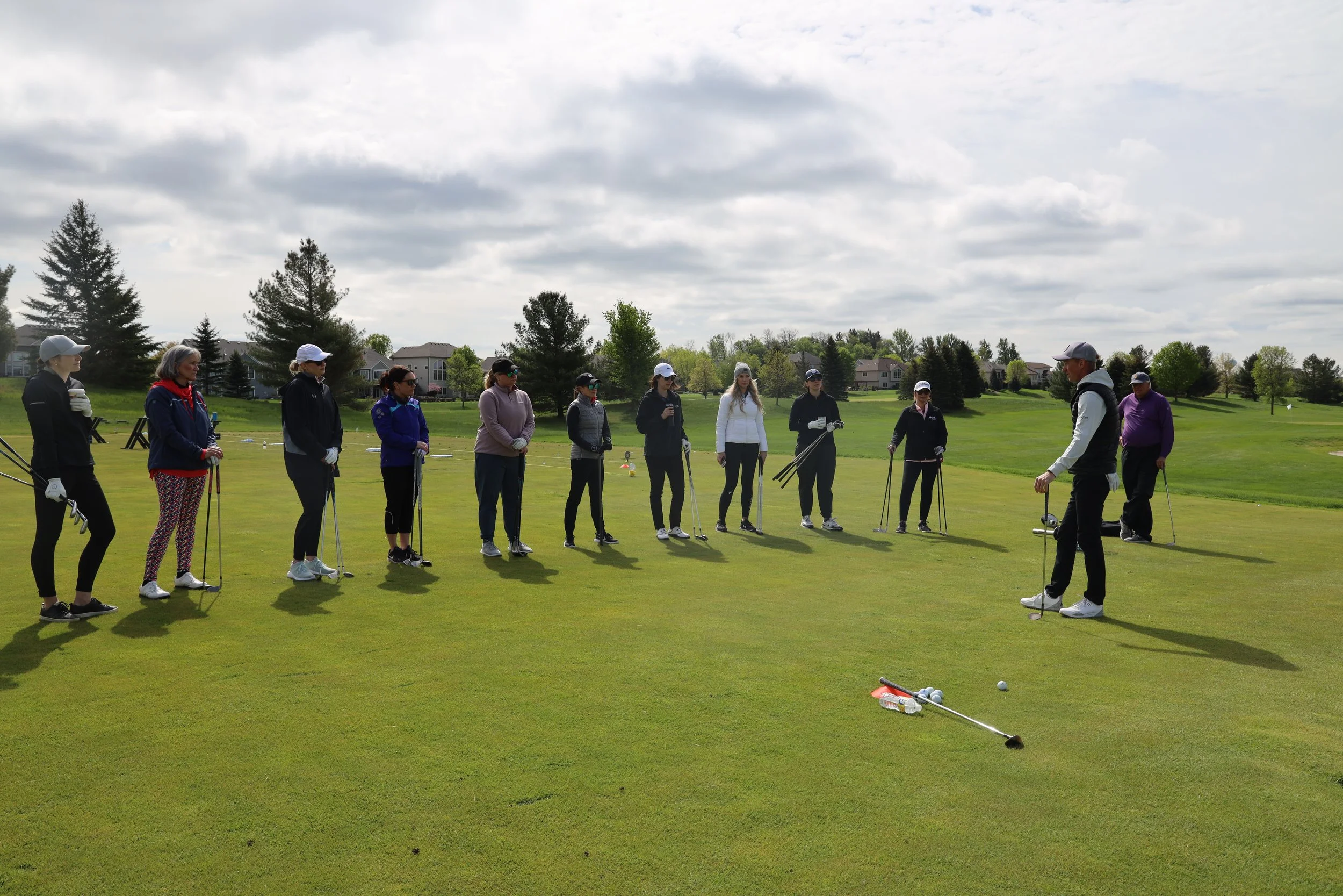 Group of people on a golf course participating in a golf lesson, with instructor demonstrating technique. Overcast skies with trees and residential houses in the background.