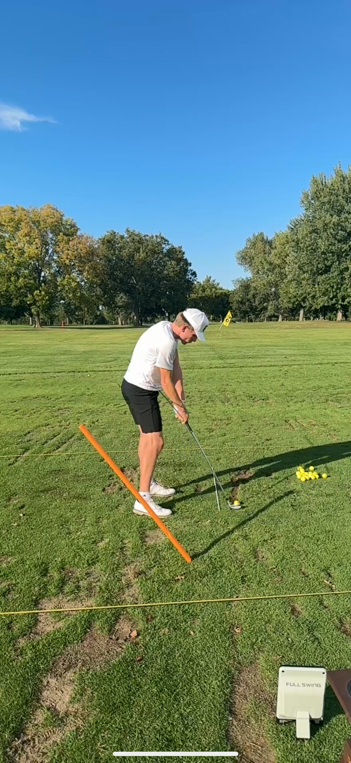 A man practicing golf on a fairway, preparing to hit a golf ball. There are multiple yellow golf balls on the ground and a small sign that says "FULL SWING." The background features trees and a clear blue sky.