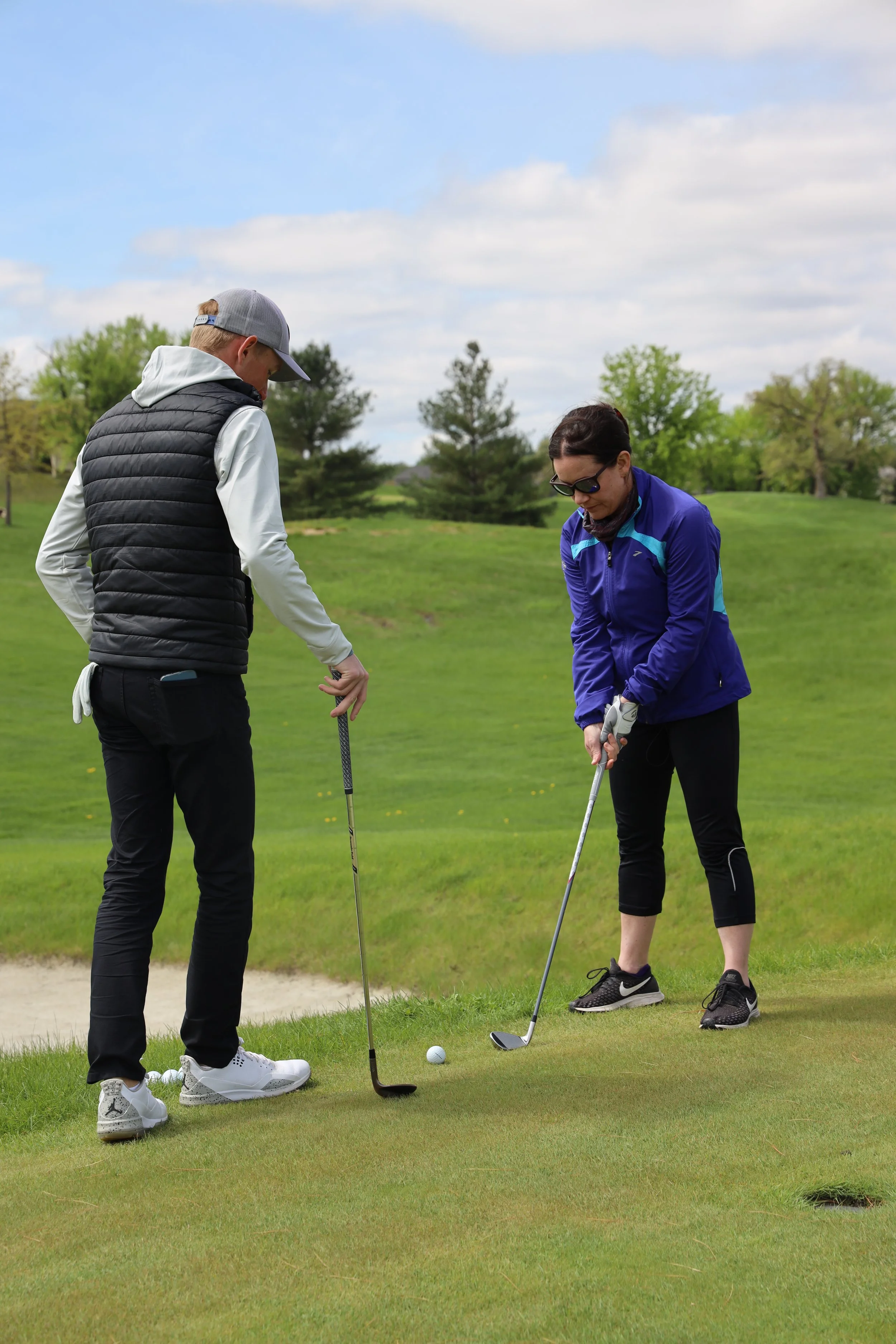 Two people on a golf course preparing for a putt, with trees and a partly cloudy sky in the background.